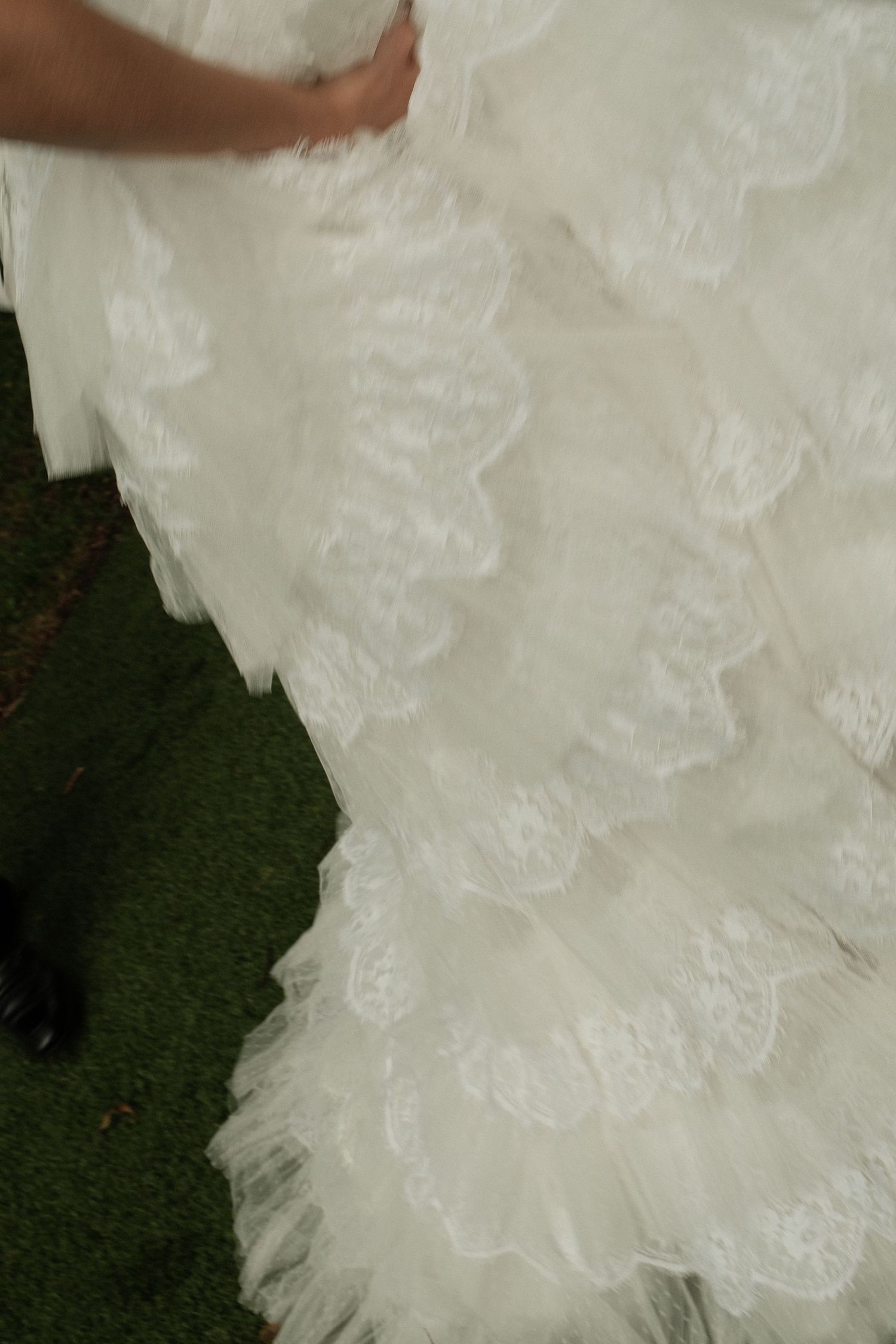 Close-up of a white lace ruffled wedding dress train on green grass, with bride and groom visible.