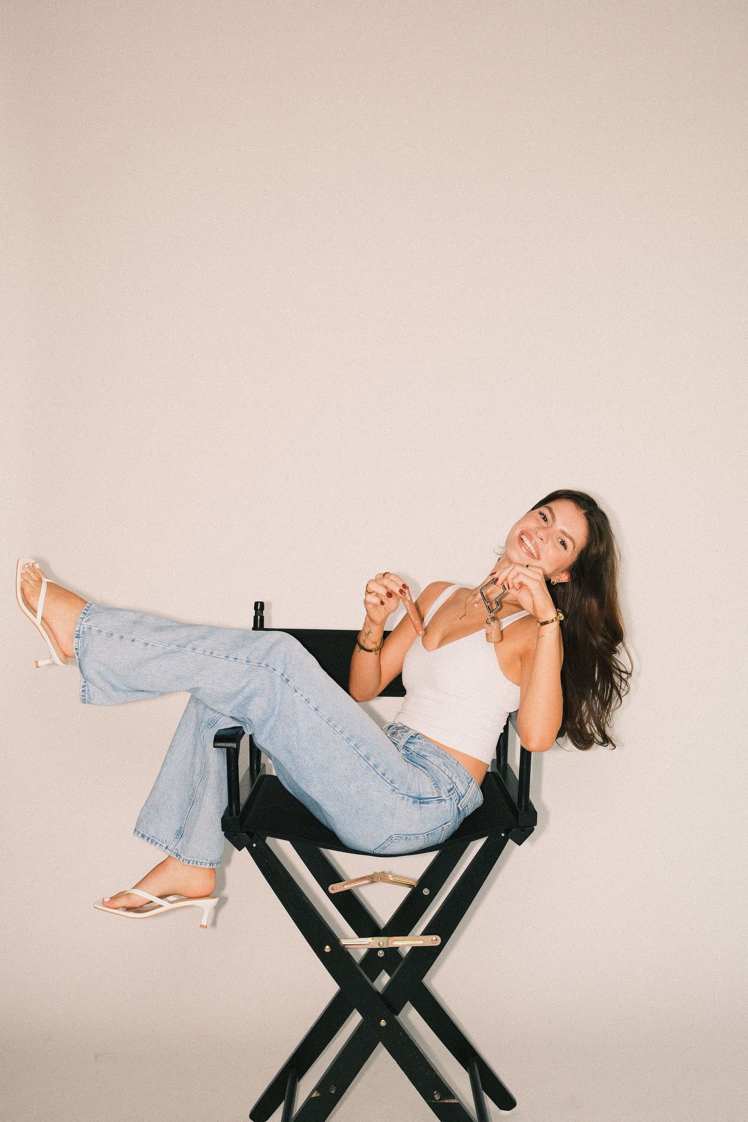 Woman in white crop top and wide-leg jeans sitting relaxed in a director's chair, smiling against a neutral background.
