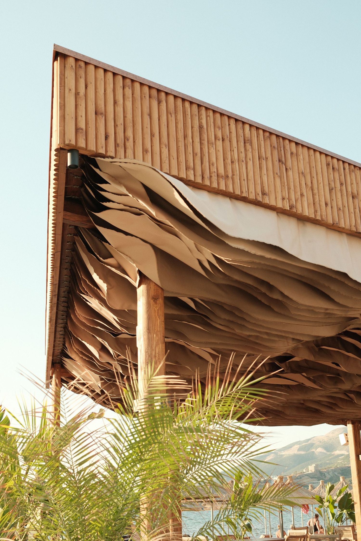 Modern architectural structure with wave-like wooden panels beneath a slatted roof, framed by tropical palm trees and mountains.