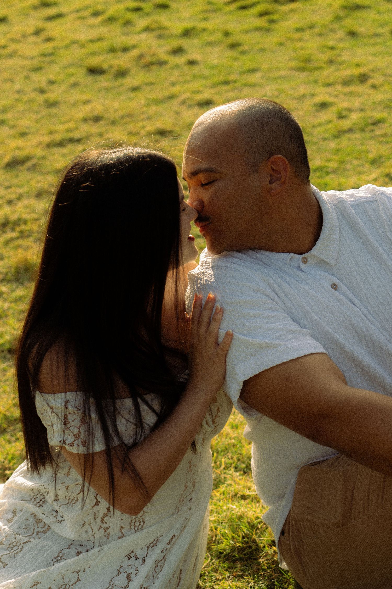 A couple shares a romantic kiss outdoors on a sunny green hillside, woman in white lace dress.