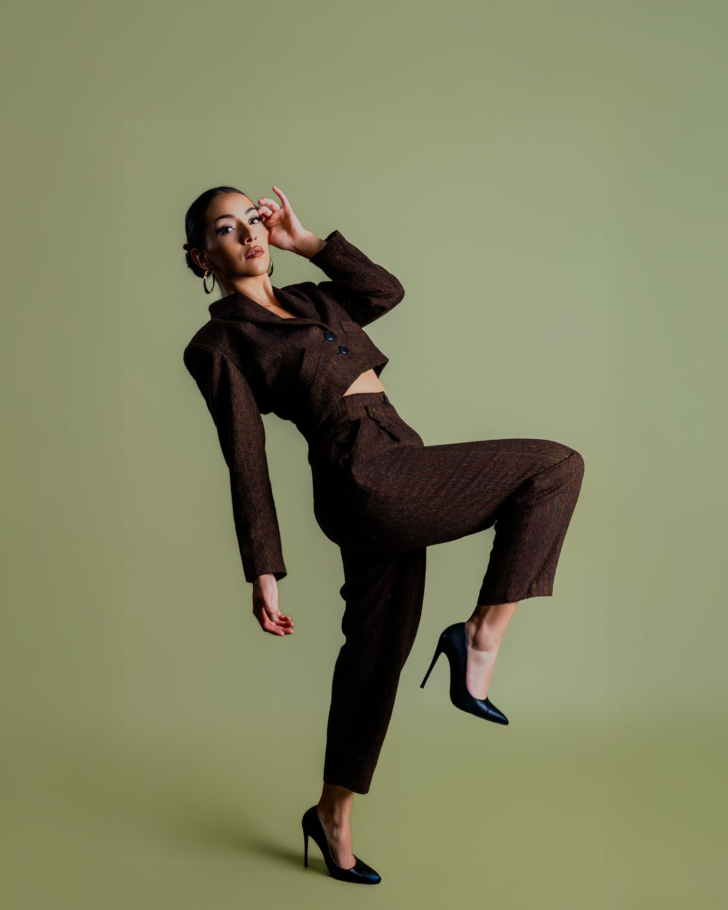 Model in brown pantsuit strikes dynamic pose with raised leg against olive green background.