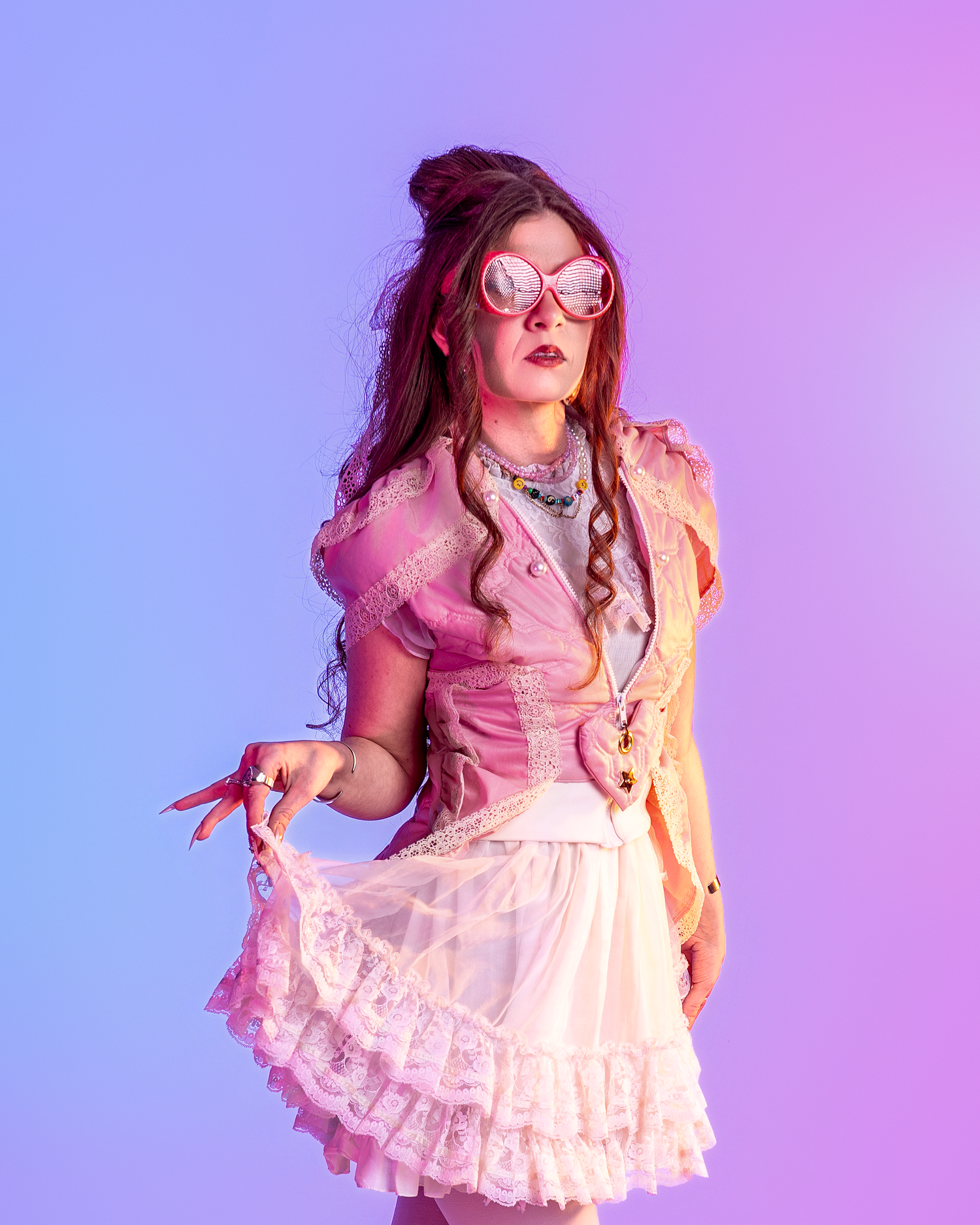 Model in pink lace dress and glasses poses playfully against purple gradient background.