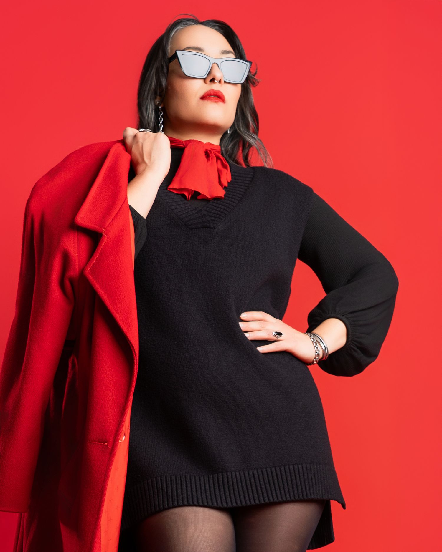 Fashion poses in black sweater and red coat against bright red background.