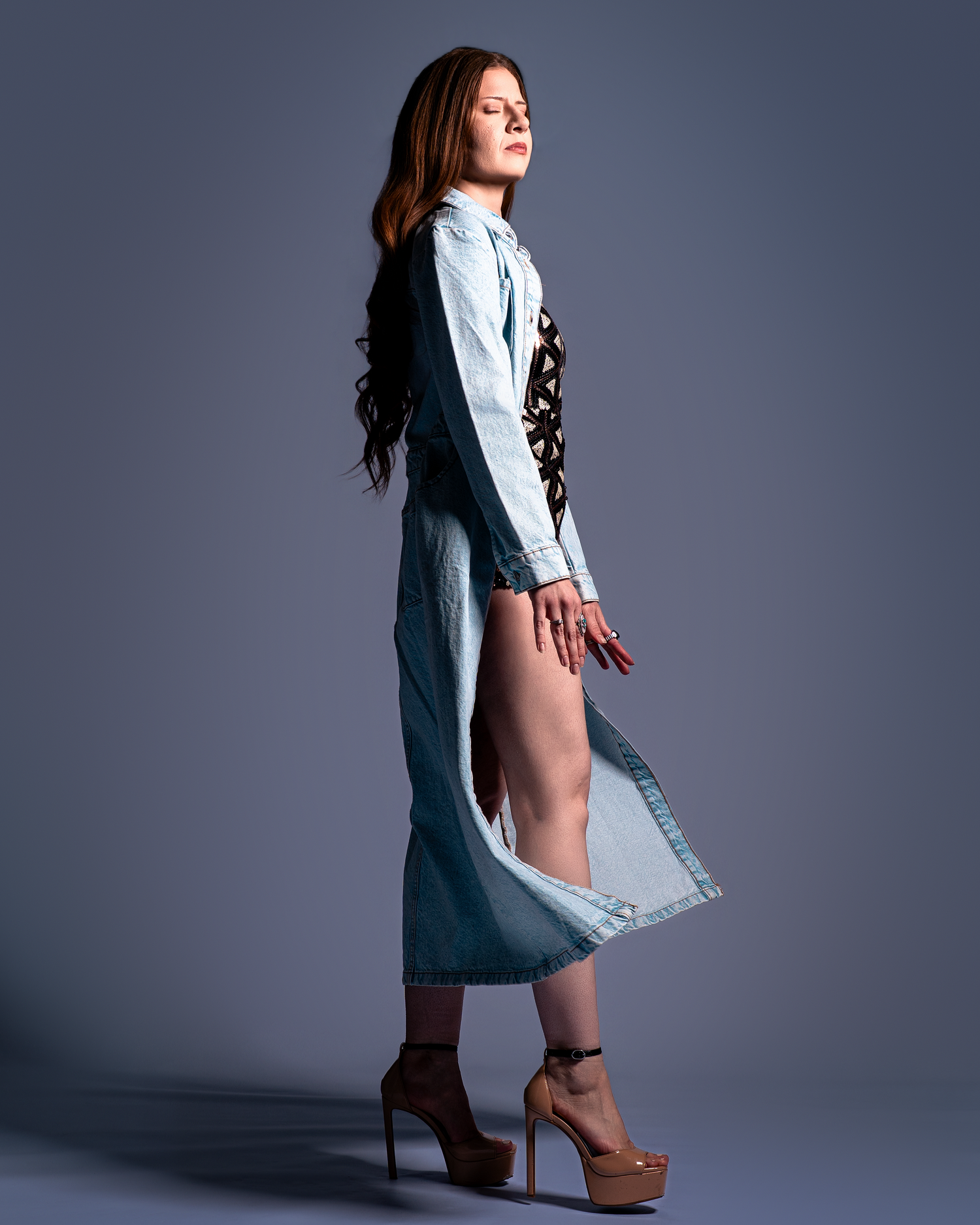 Fashion model in denim dress and platform heels poses against gray backdrop in dramatic studio lighting.