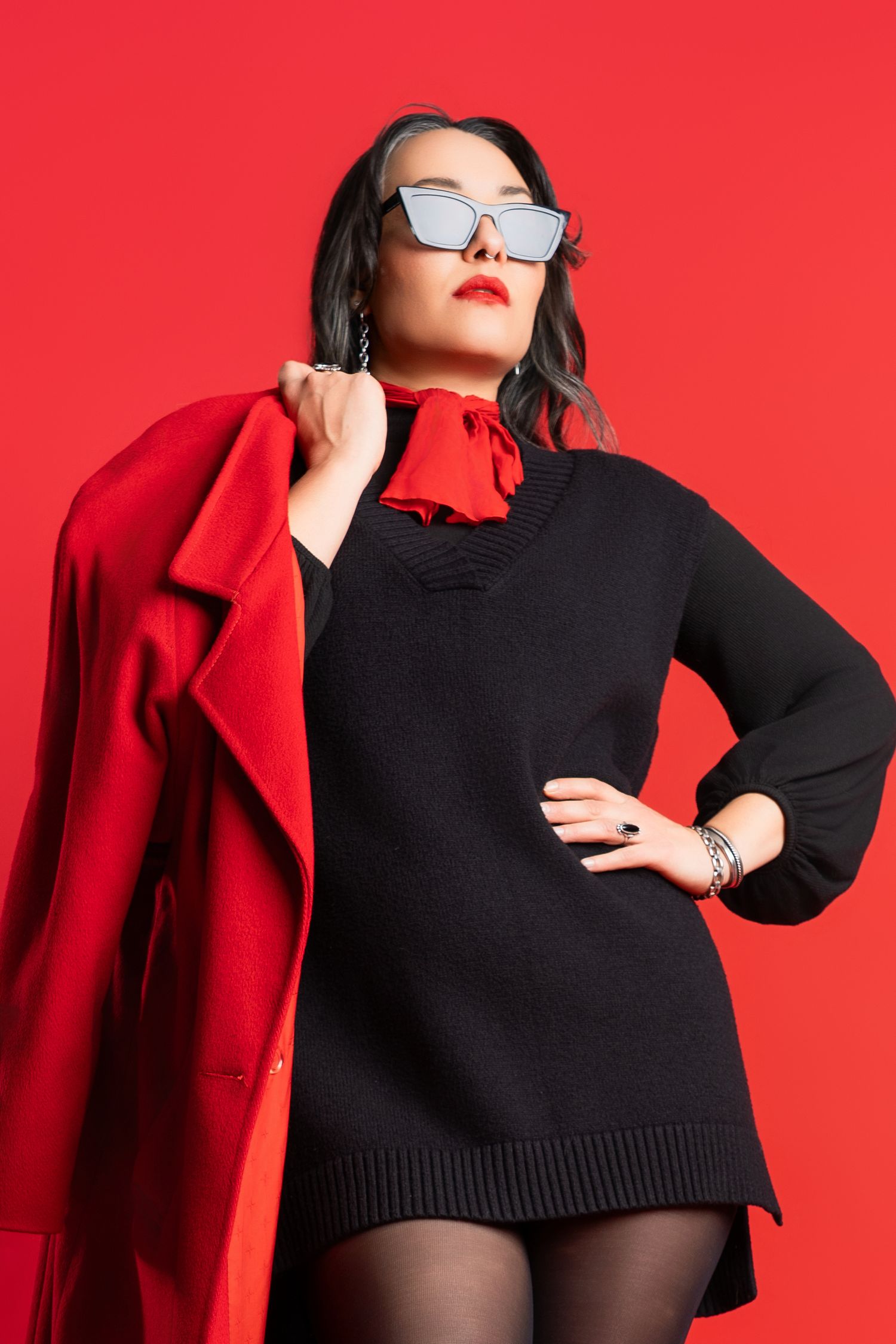 Fashion model wearing black sweater and red coat poses against vibrant red background.