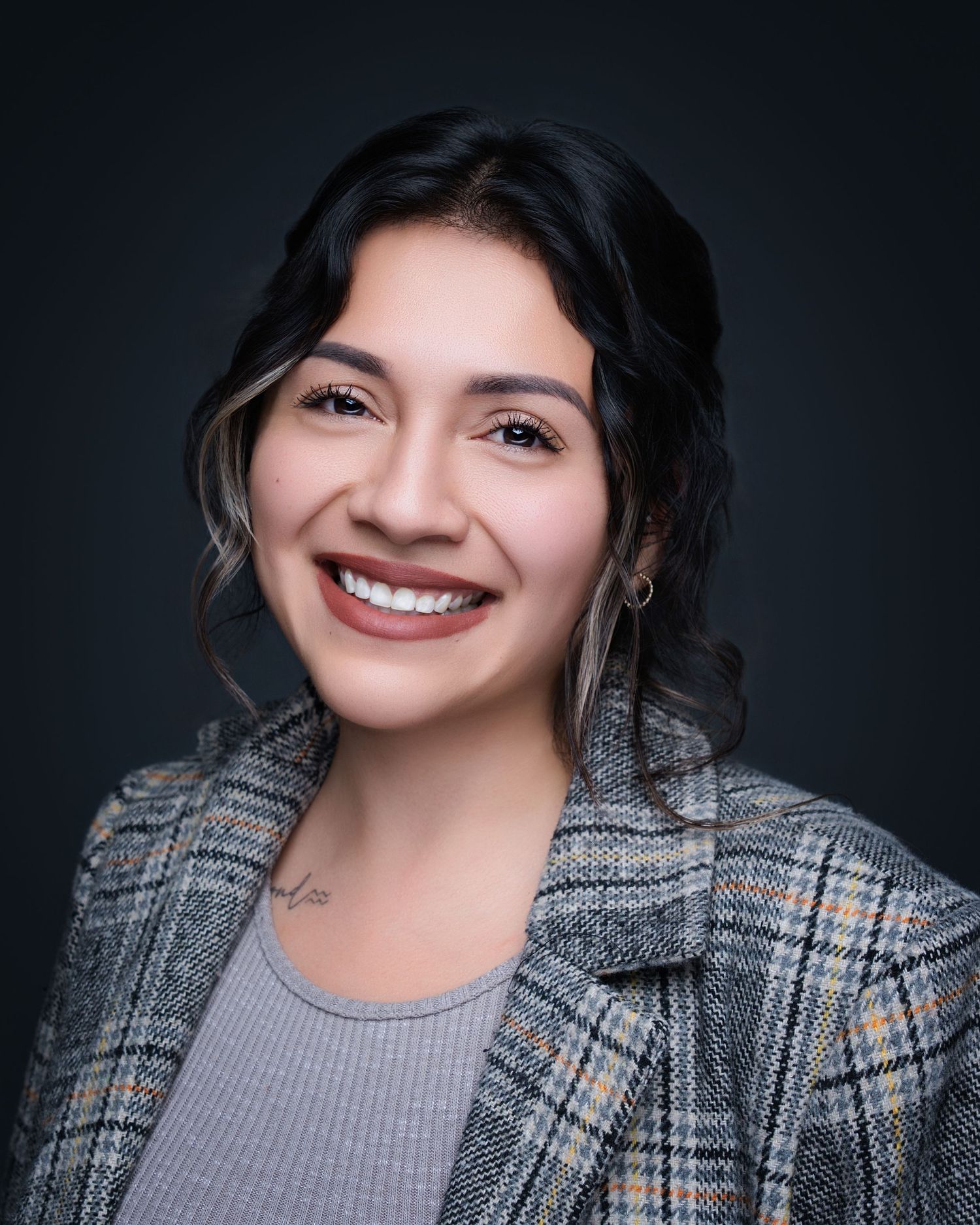 Professional headshot portrait in grey sweater and plaid blazer against dark background with warm smile.