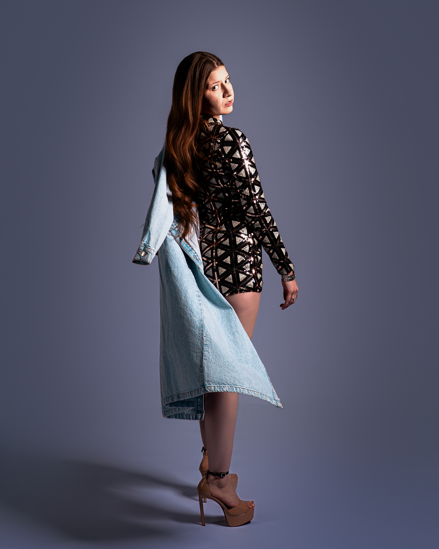 A model poses in a patterned mini dress and denim coat against a gray backdrop while wearing platform heels.
