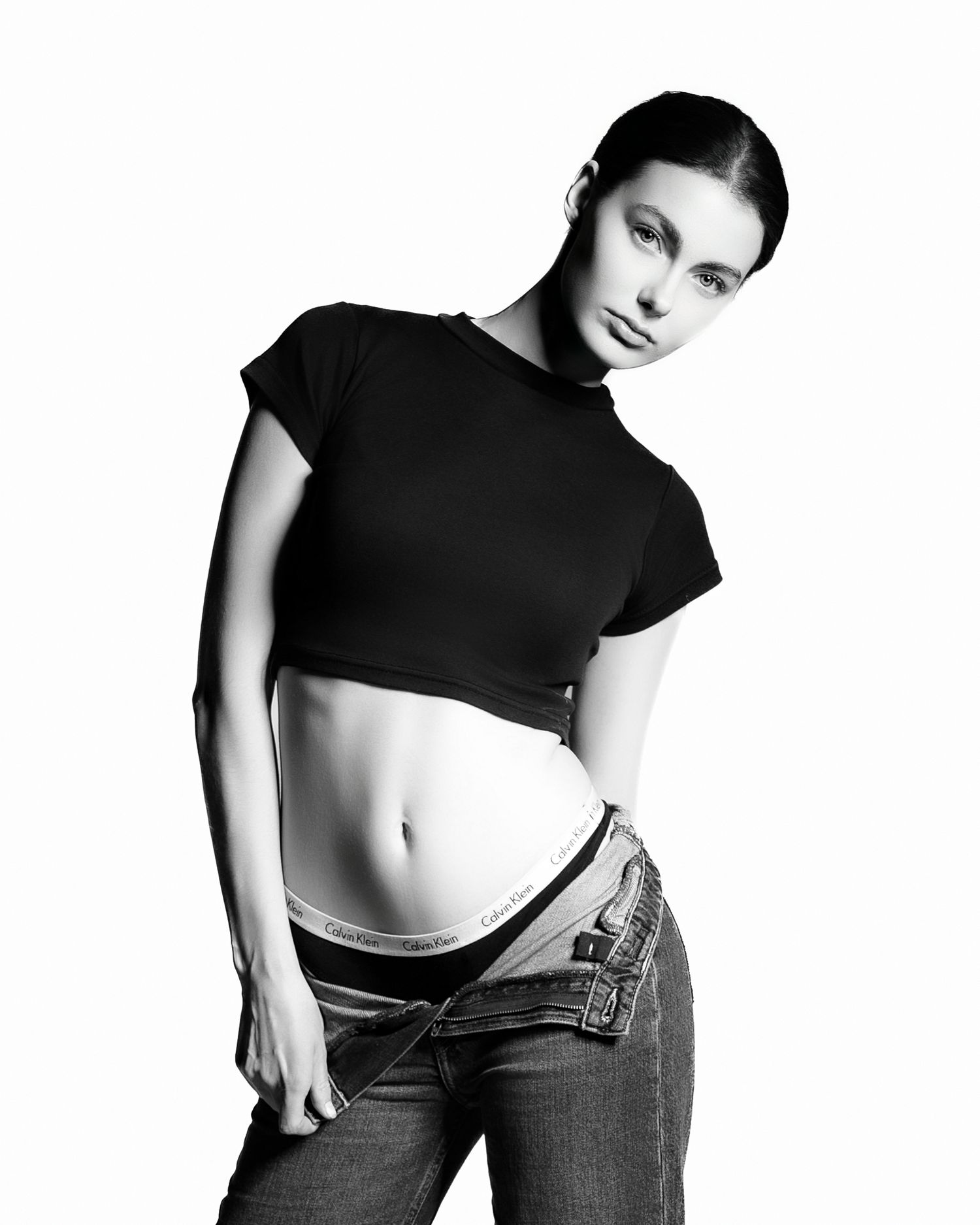 Black and white fashion photo of a model in cropped black top and jeans posing against white background.