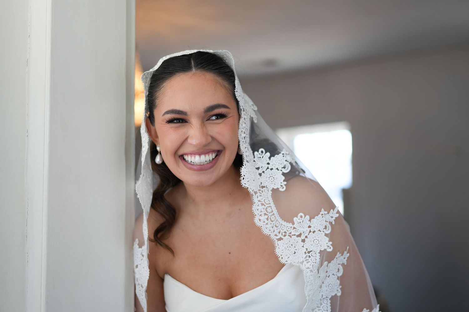 Smiling bride in white strapless gown and lace-trimmed veil posing elegantly in a bright doorway.