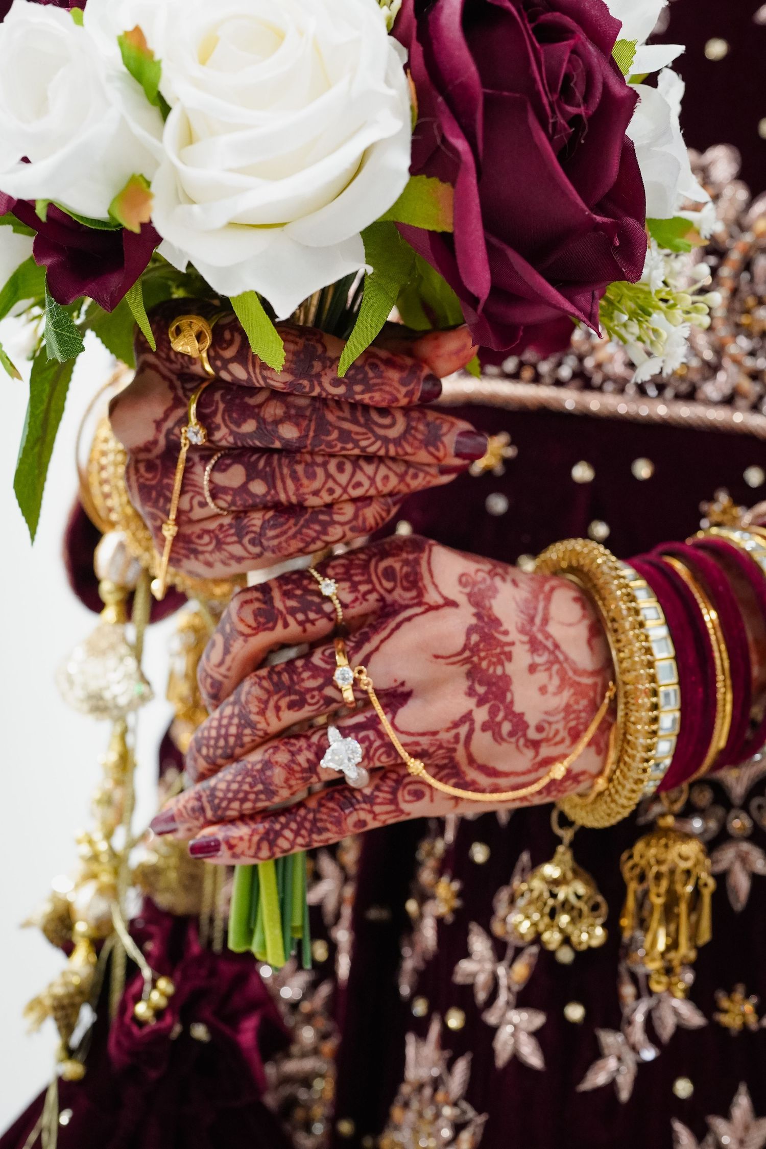 photoshoot of bride's henna-adorned hands holding a bouquet of burgundy and white roses with delicate gold bangles and jewelry.