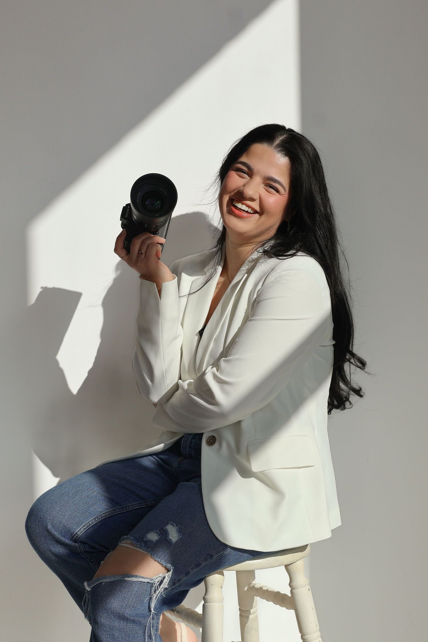 Smiling woman in white blazer and jeans holding a camera, seated on a stool against a bright white wall.