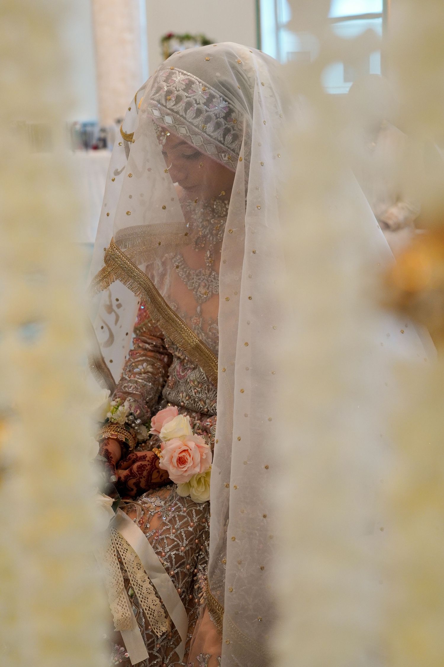 Bride in embellished veil holding pink roses in soft natural light.