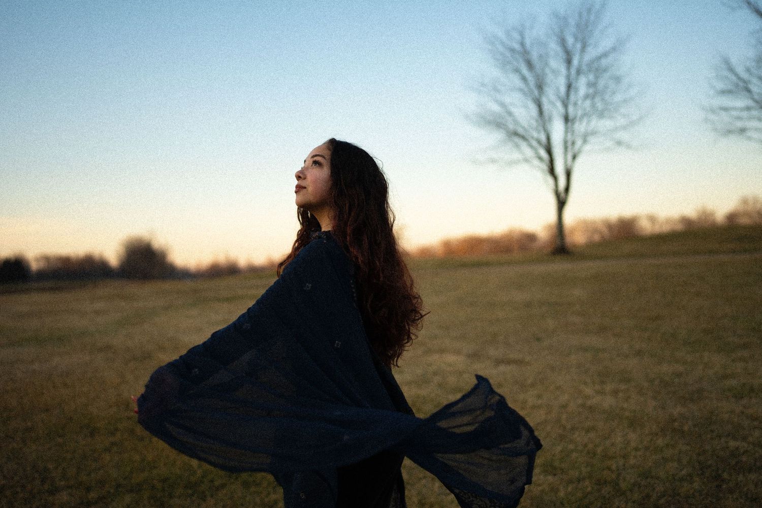 photography of a girl in a flowing black dress spins gracefully in a field at sunset creating a dramatic silhouette effect.