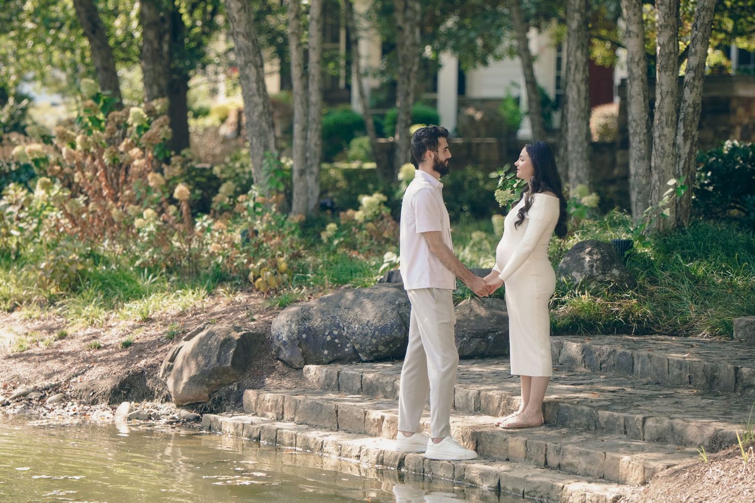 A couple in elegant white attire poses together in a serene outdoor setting with stone walls and lush greenery.
