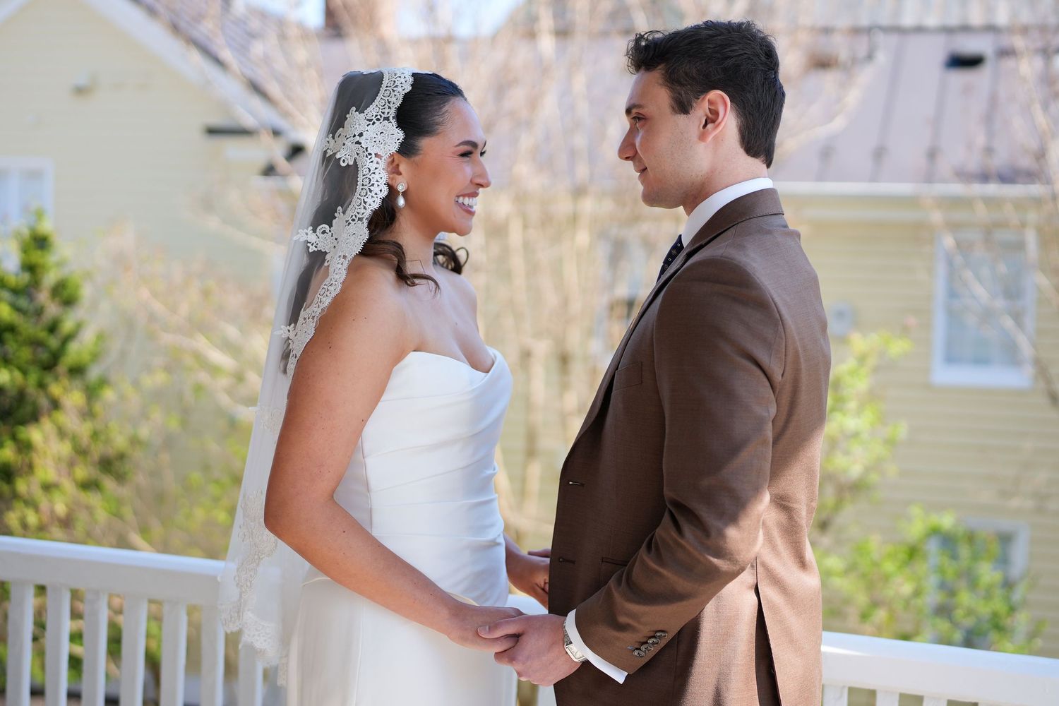 Bride in white gown and veil holds hands with groom in brown suit on a decorated outdoor balcony.