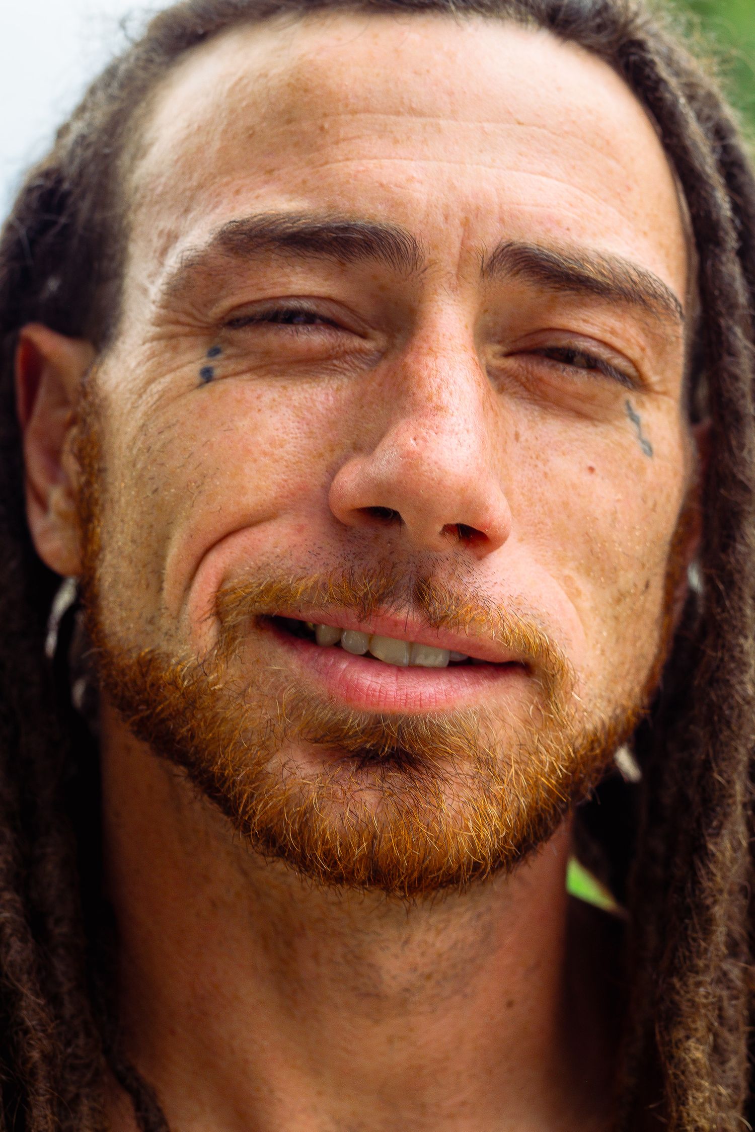 Close-up portrait of smiling man with dreadlocks, red beard, and small facial tattoos.