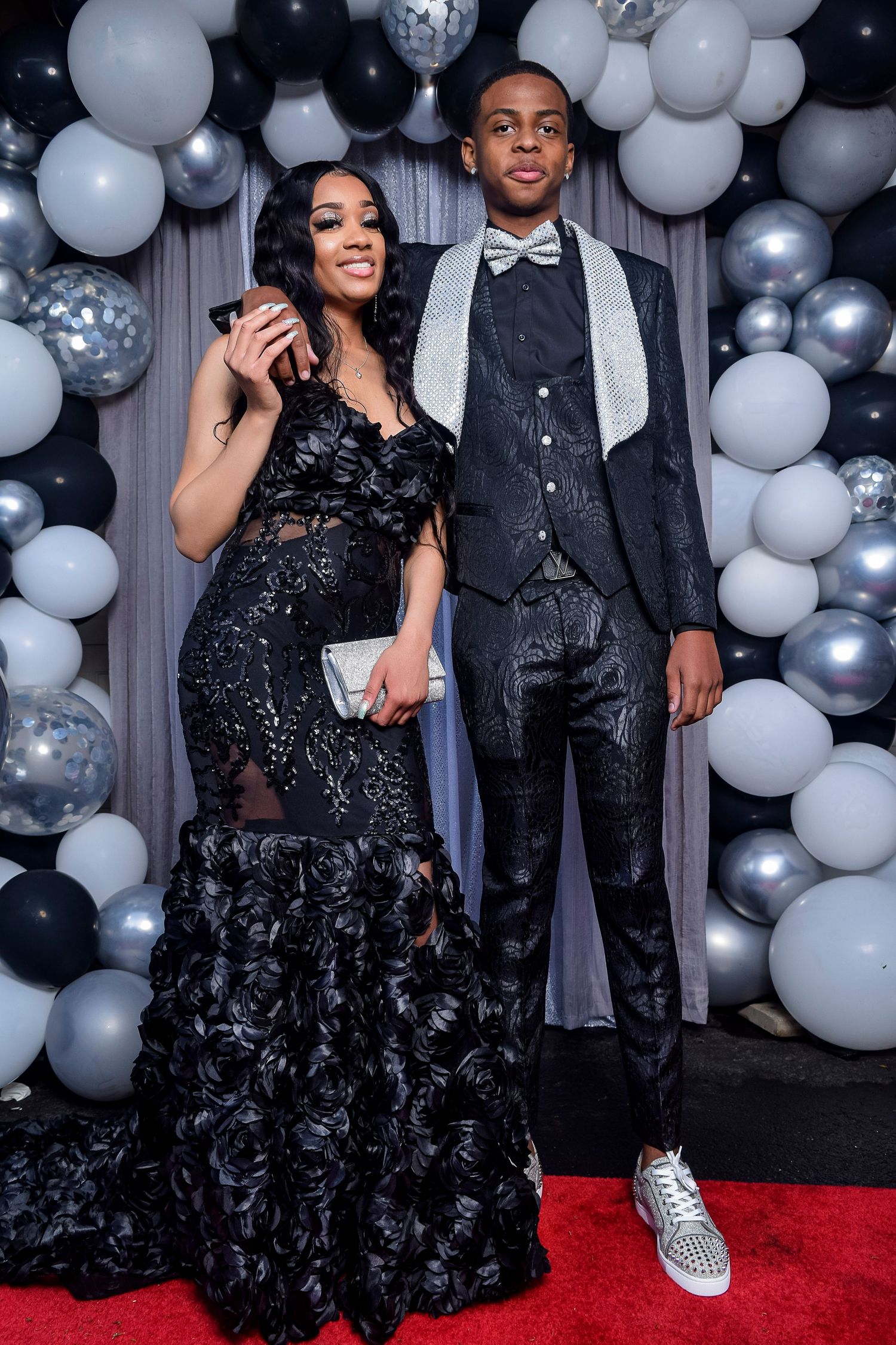 Couple posing at prom in matching black outfits with balloon backdrop.