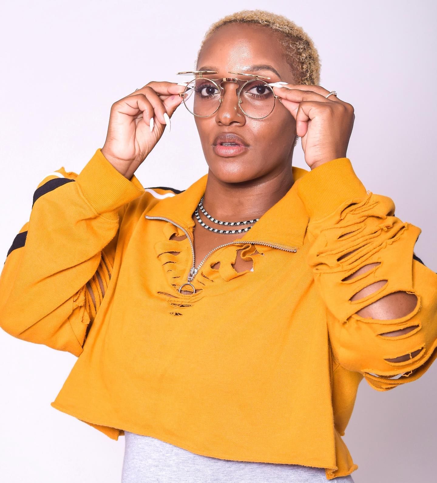 Woman in mustard yellow distressed sweatshirt adjusting round wire-rimmed glasses against white background.