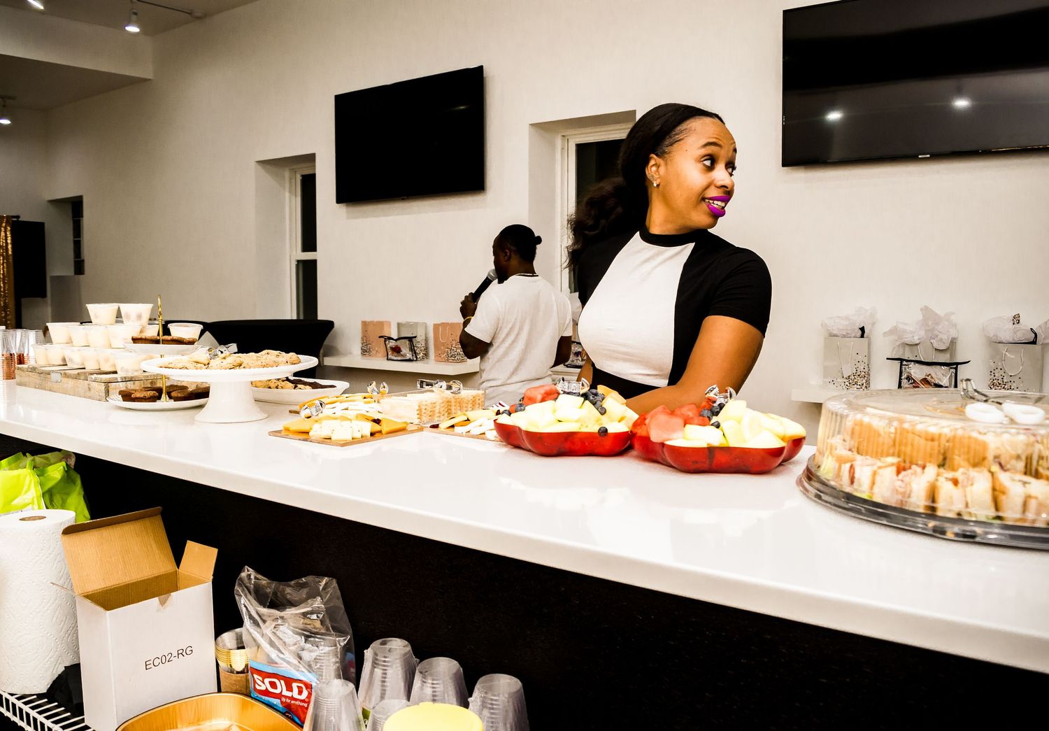 Woman in black and white outfit stands behind catered food spread at indoor event.