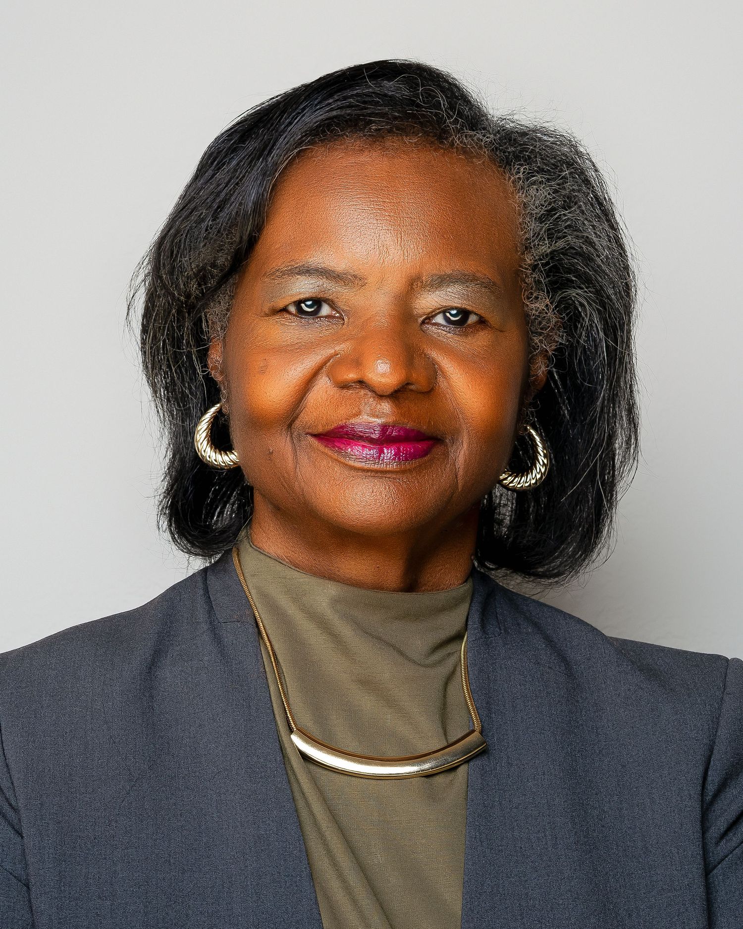 Professional headshot of a mature woman in a gray blazer with gold hoop earrings and necklace.