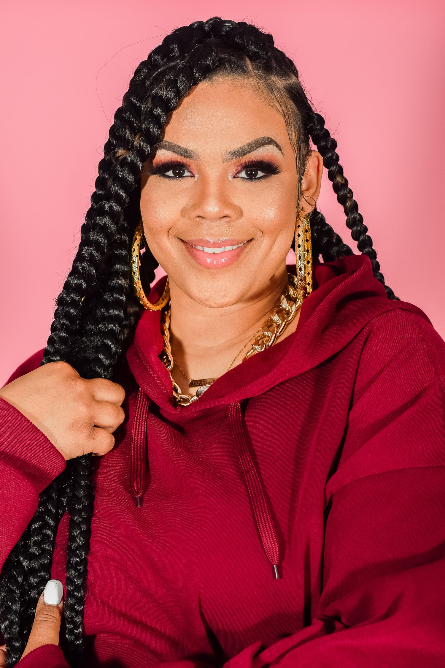 Smiling woman in burgundy hoodie with long box braids and gold hoop earrings on pink background.