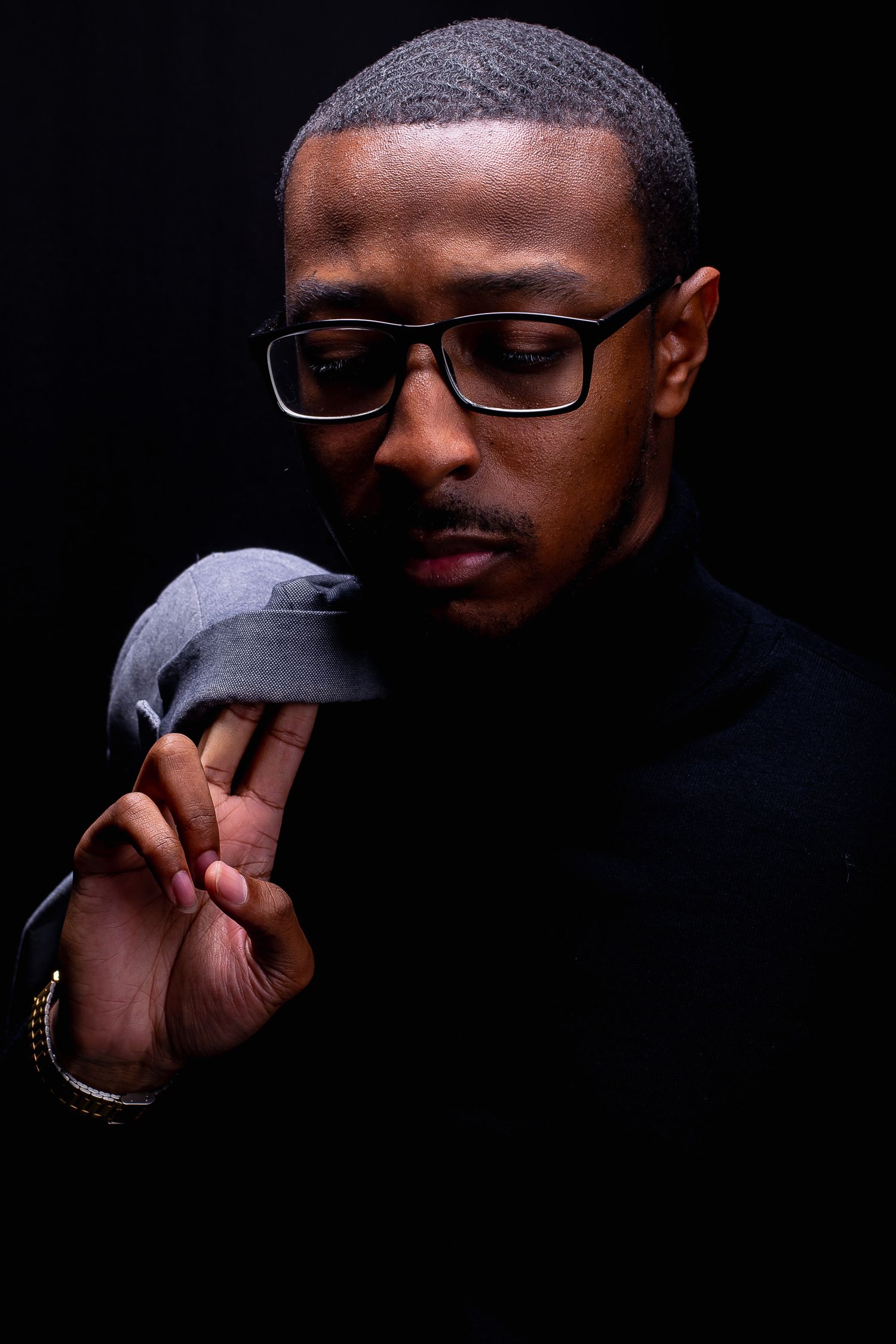Stylish Black man in black turtleneck and glasses holding a gray cap against dark background.
