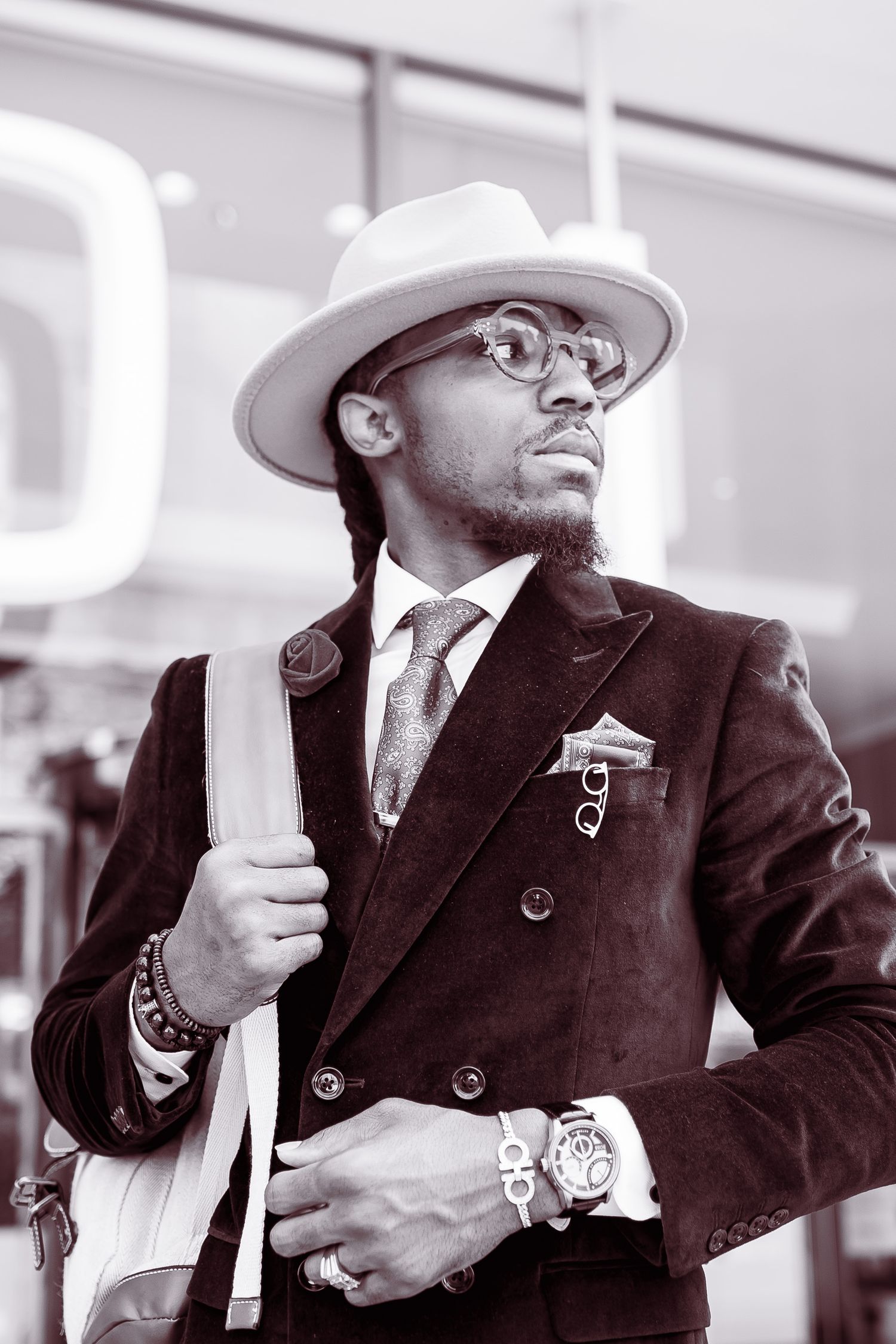 Stylish man in a white fedora, glasses, and velvet blazer posing outdoors in a black and white photo.