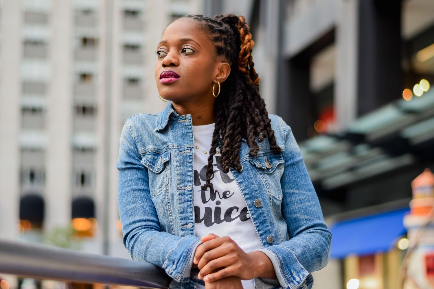 Woman in denim jacket leaning on railing outdoors, wearing white graphic tee with braided hair in city.