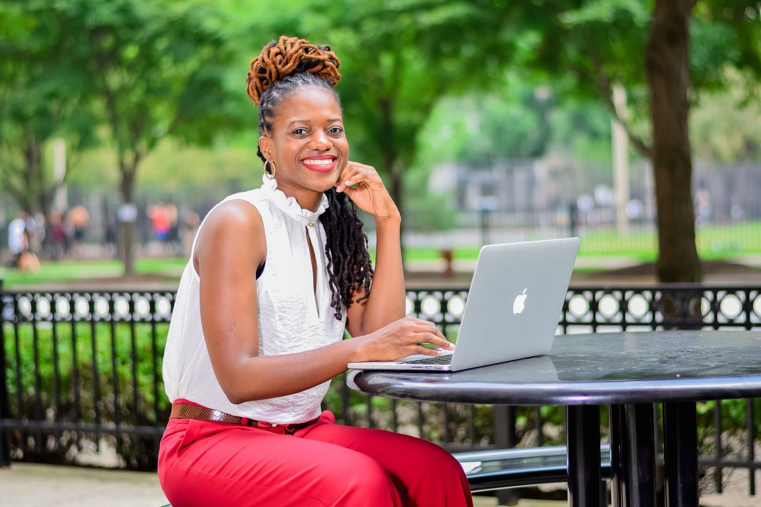Smiling woman with braided hair in white top and red pants using MacBook laptop at outdoor park table.