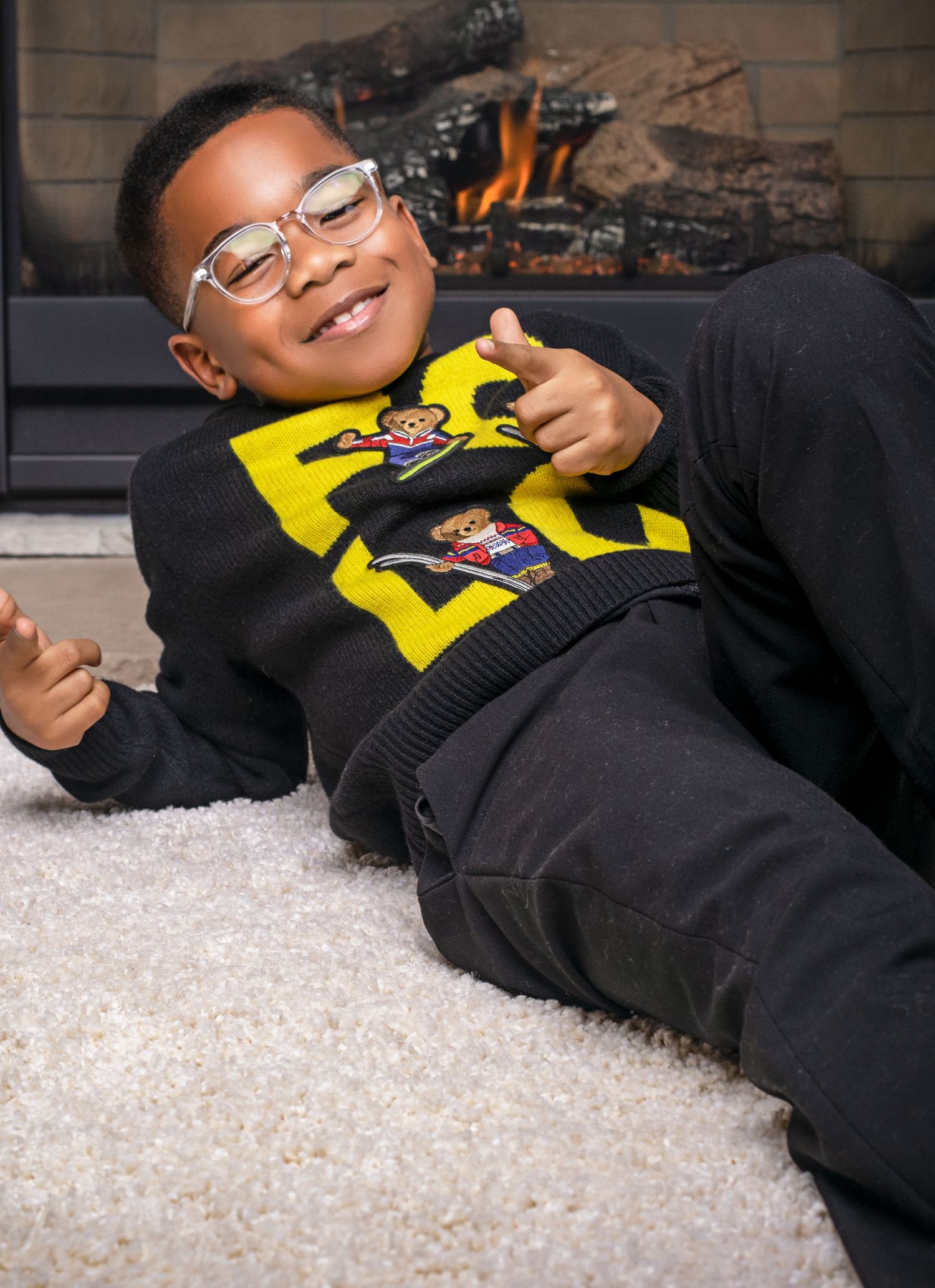 Smiling boy in yellow bear sweater poses on carpet in front of fireplace.