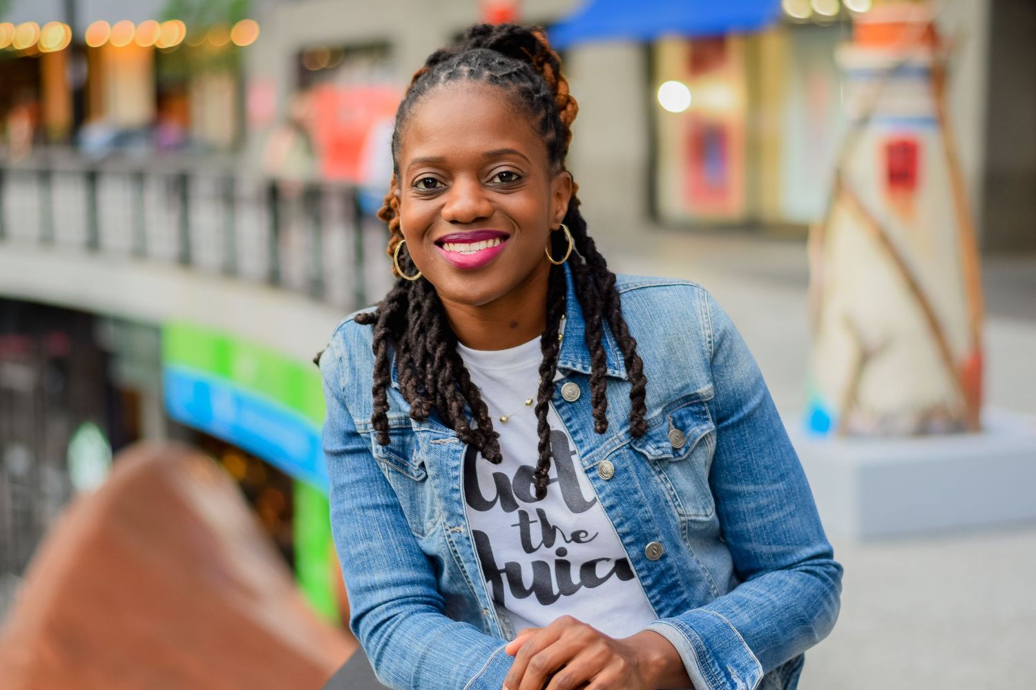 Smiling woman in denim jacket and white graphic tee with braided hair, seated outdoors in urban setting.