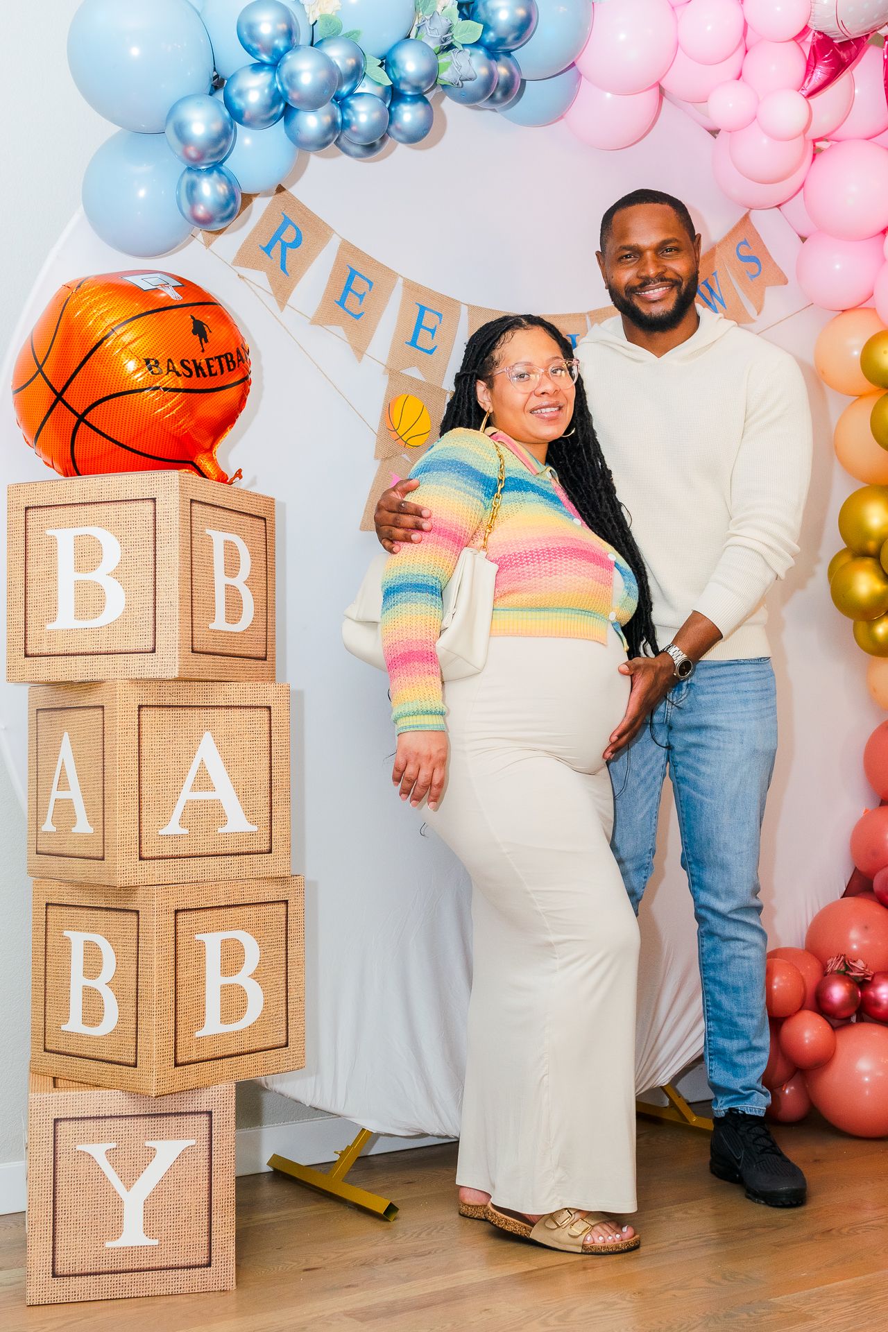 Expecting Black couple posing at basketball-themed baby shower beside stacked BABY letter blocks.