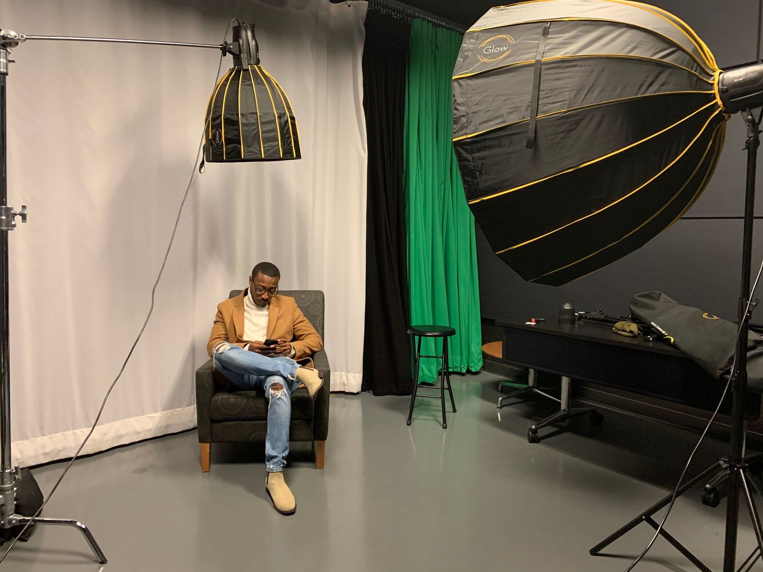 Man sitting in armchair in photography studio with green screen backdrop and professional lighting equipment.