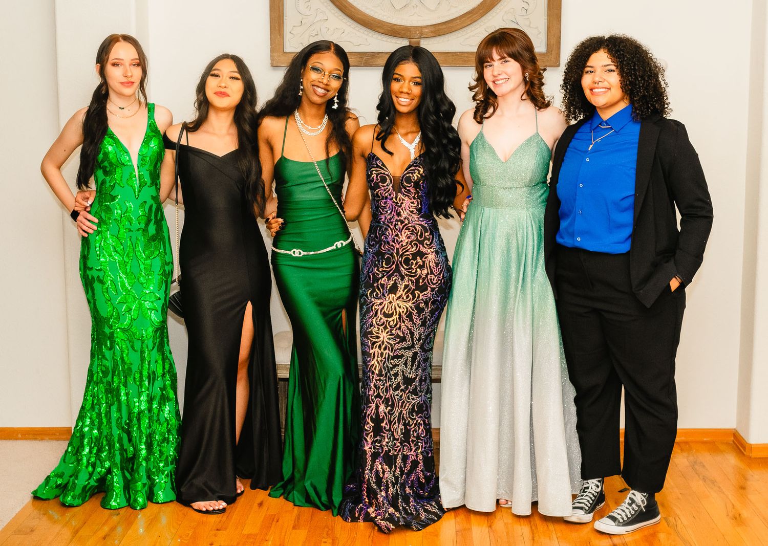 Six young women in elegant formal gowns posing together at a prom or formal event.