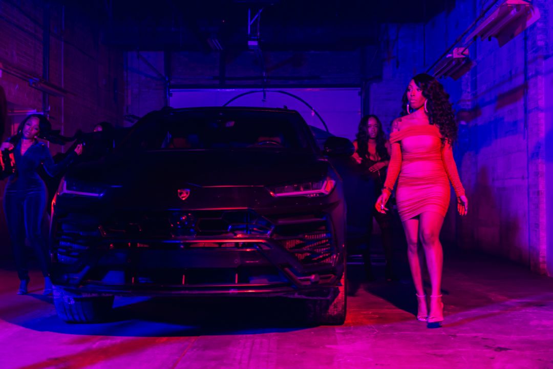 A woman in a pink outfit walks past a black luxury car under purple neon lights.