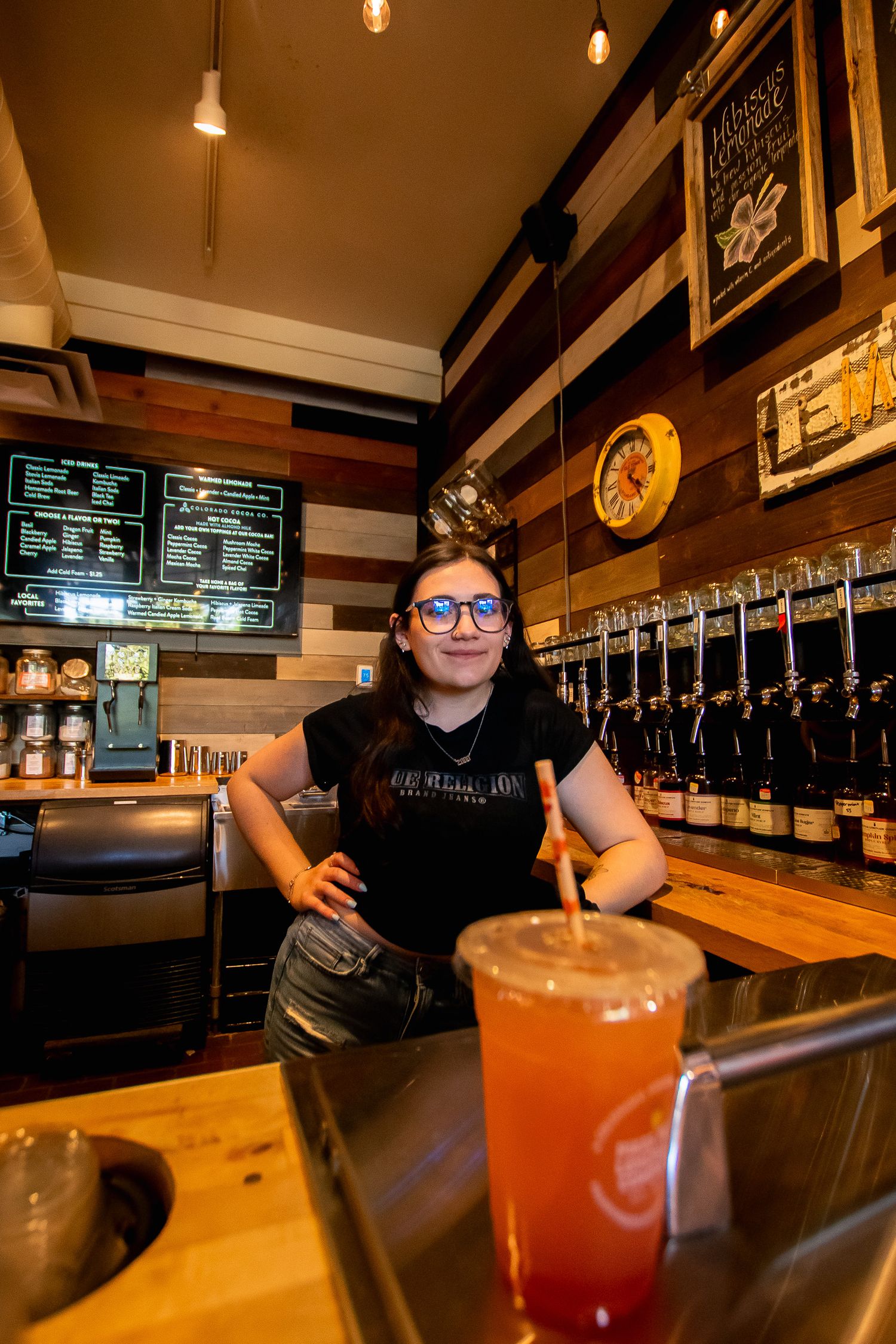 A smiling female bartender poses behind a wooden bar counter with a colorful drink.