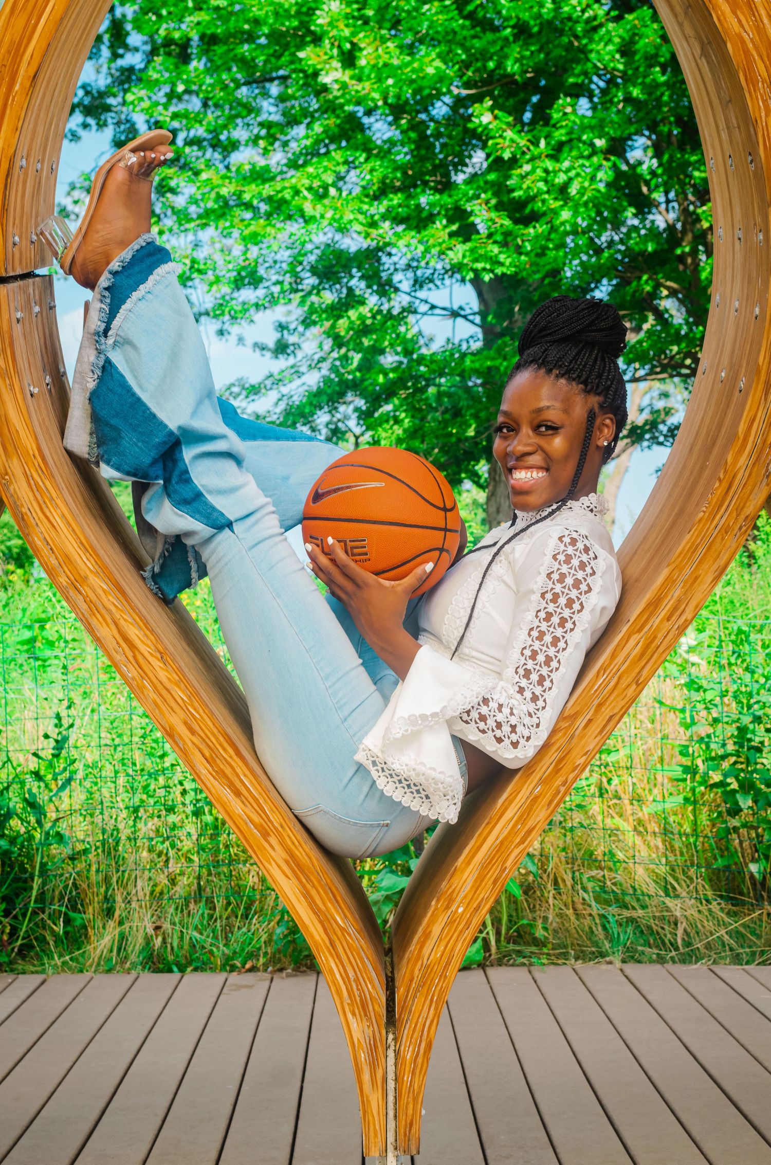 Smiling young Black woman holding basketball while sitting inside heart-shaped wooden outdoor sculpture.