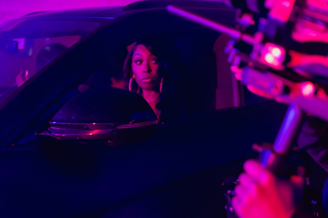 A woman sits in a dark car illuminated by purple and pink neon lights on a film set.