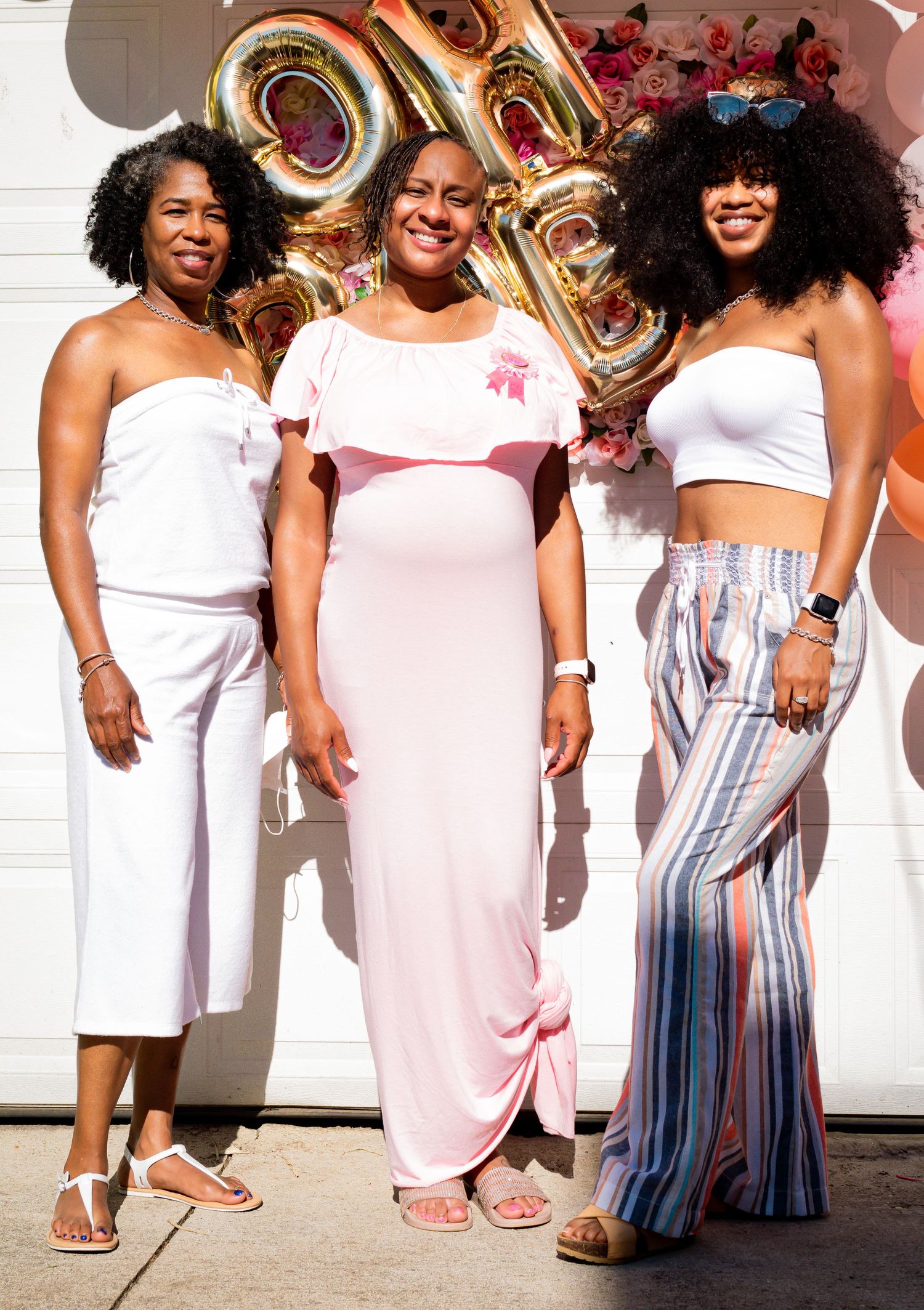 Three women posing together at an outdoor baby shower with gold OH BABY balloon letters behind them.