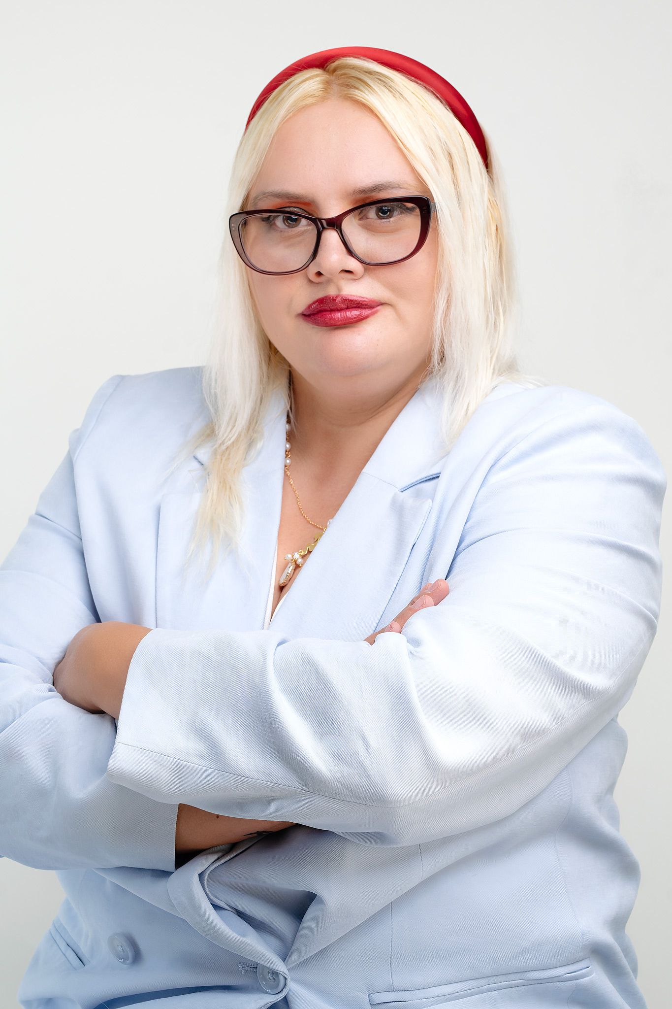 Confident blonde woman with glasses and red headband poses arms crossed in light blue blazer.