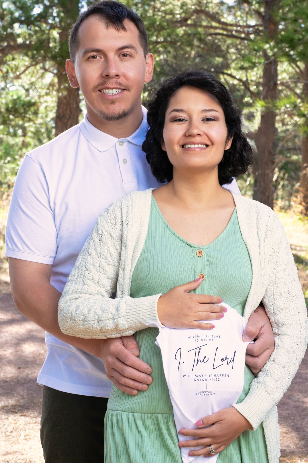Smiling couple posing outdoors with a pregnant woman holding a Christian baby onesie.
