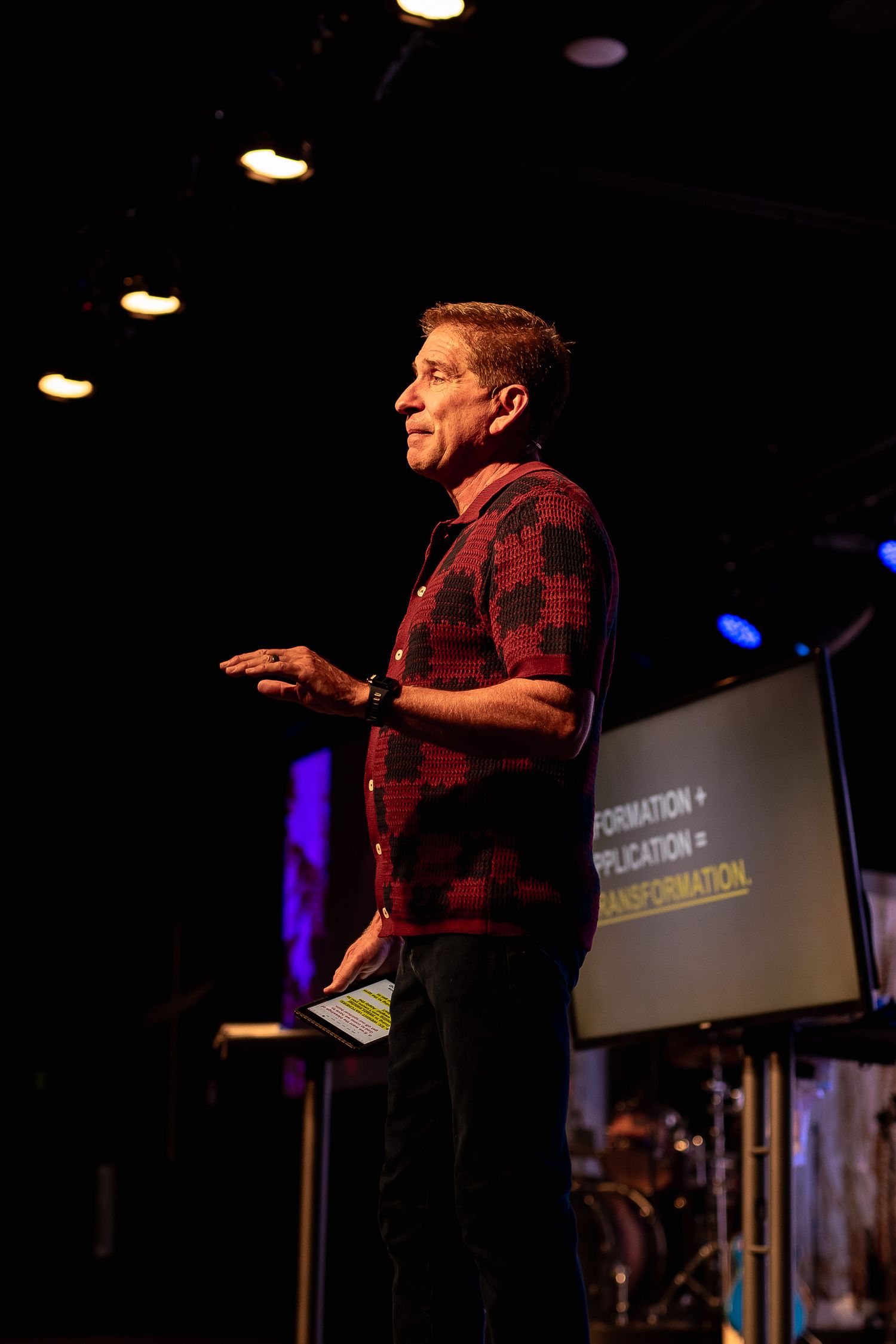 Man in a red floral shirt speaking on a dimly lit stage with a presentation screen behind him.