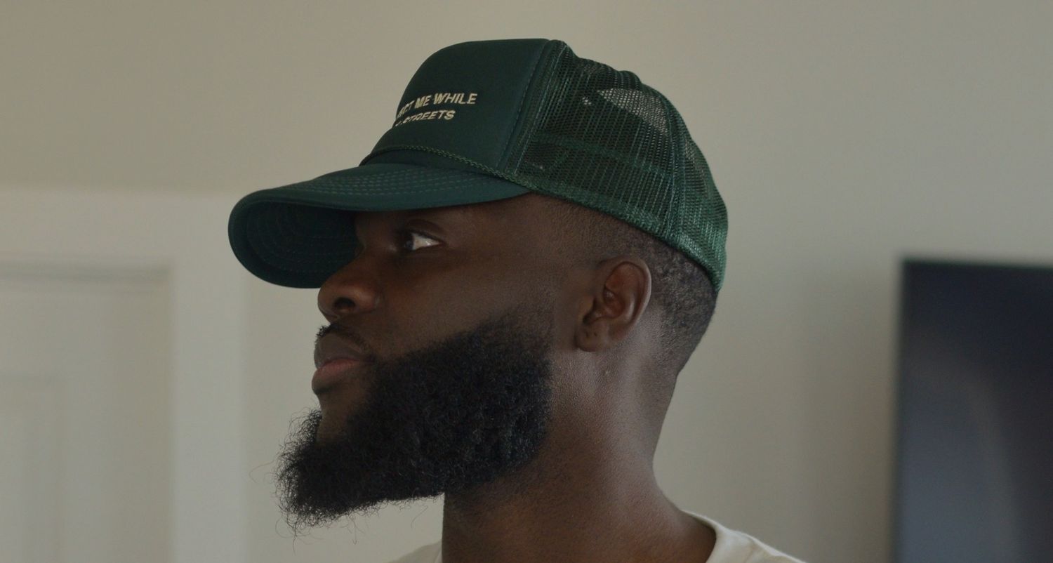 Side profile of a bearded Black man wearing a green trucker cap in a softly lit room.
