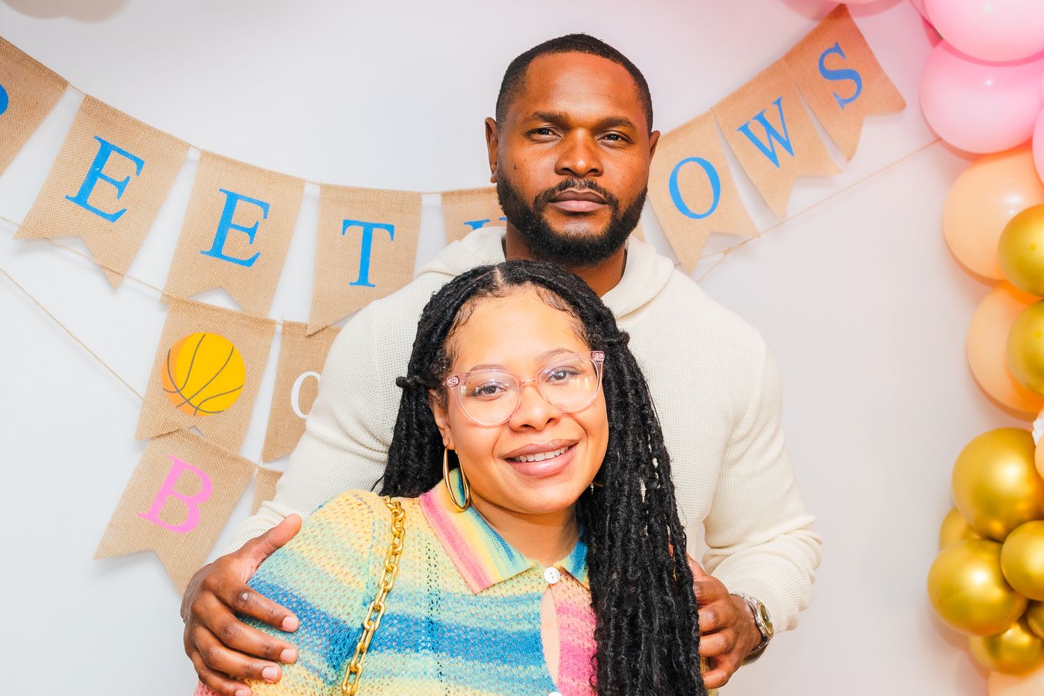 Smiling Black couple posing together at a baby shower with burlap banner and balloon decorations.