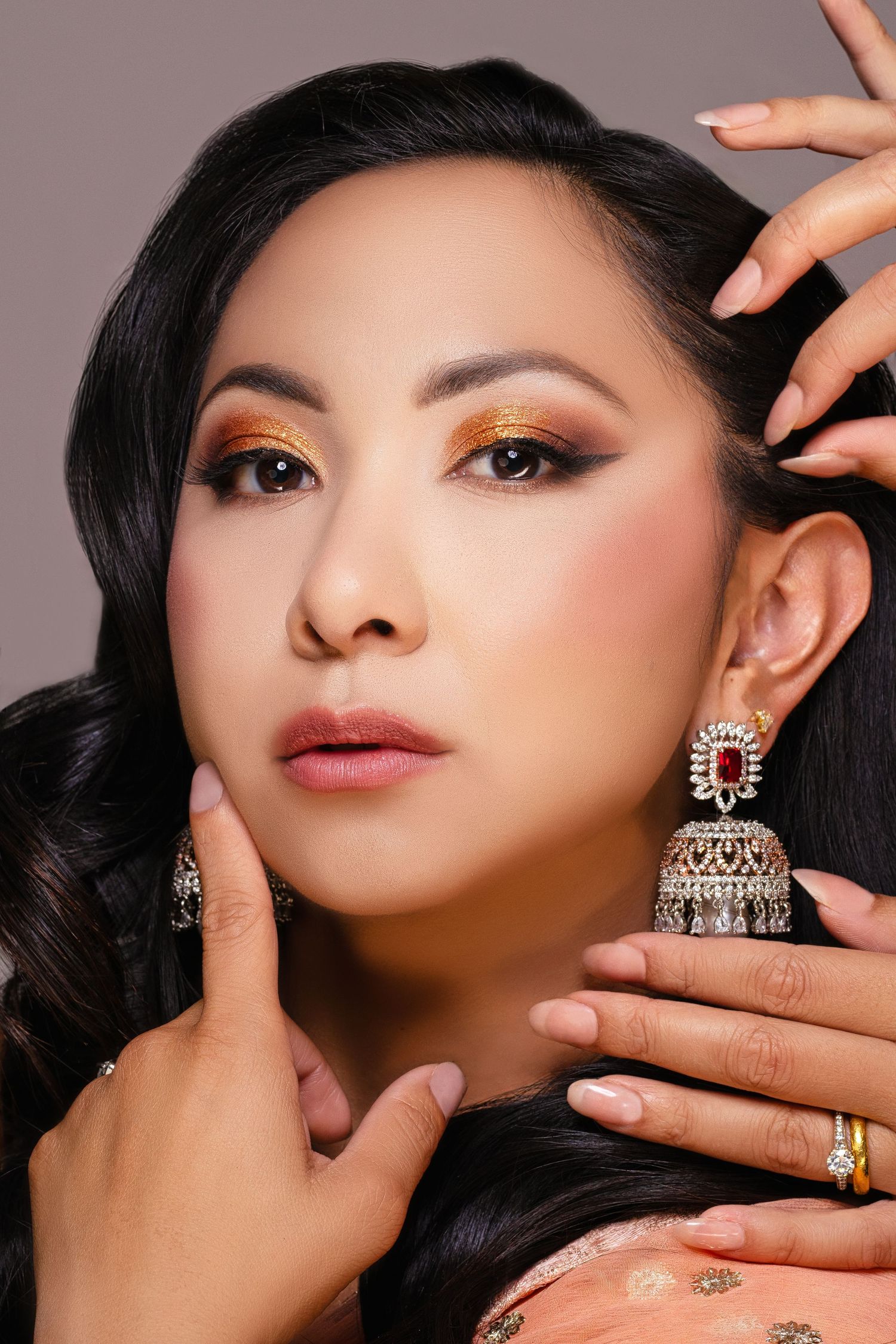 Asian woman with golden smoky eye makeup and ornate silver jhumka earrings poses gracefully on gray background.