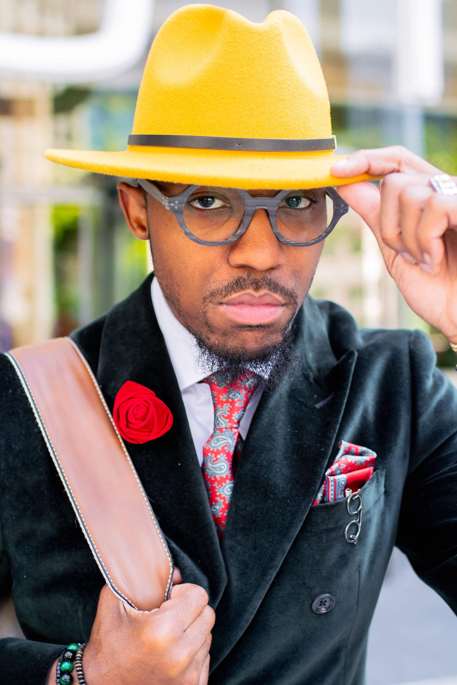 Stylish Black man in yellow fedora, gray glasses, and black velvet blazer with red rose pin.