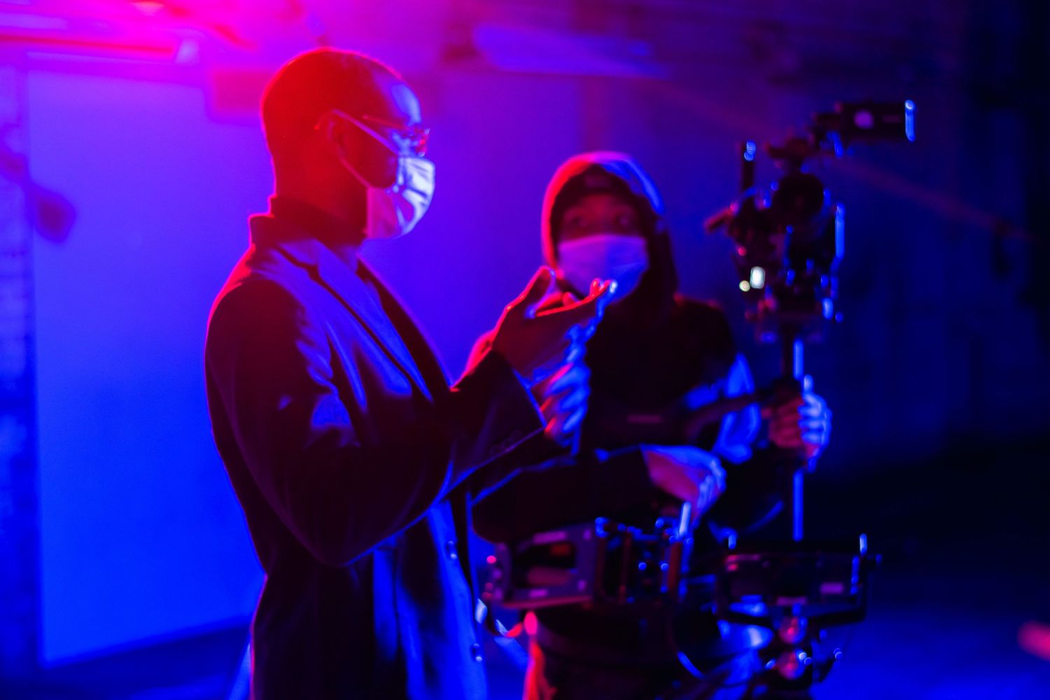 Film crew wearing masks operate camera equipment under dramatic blue and red lighting.