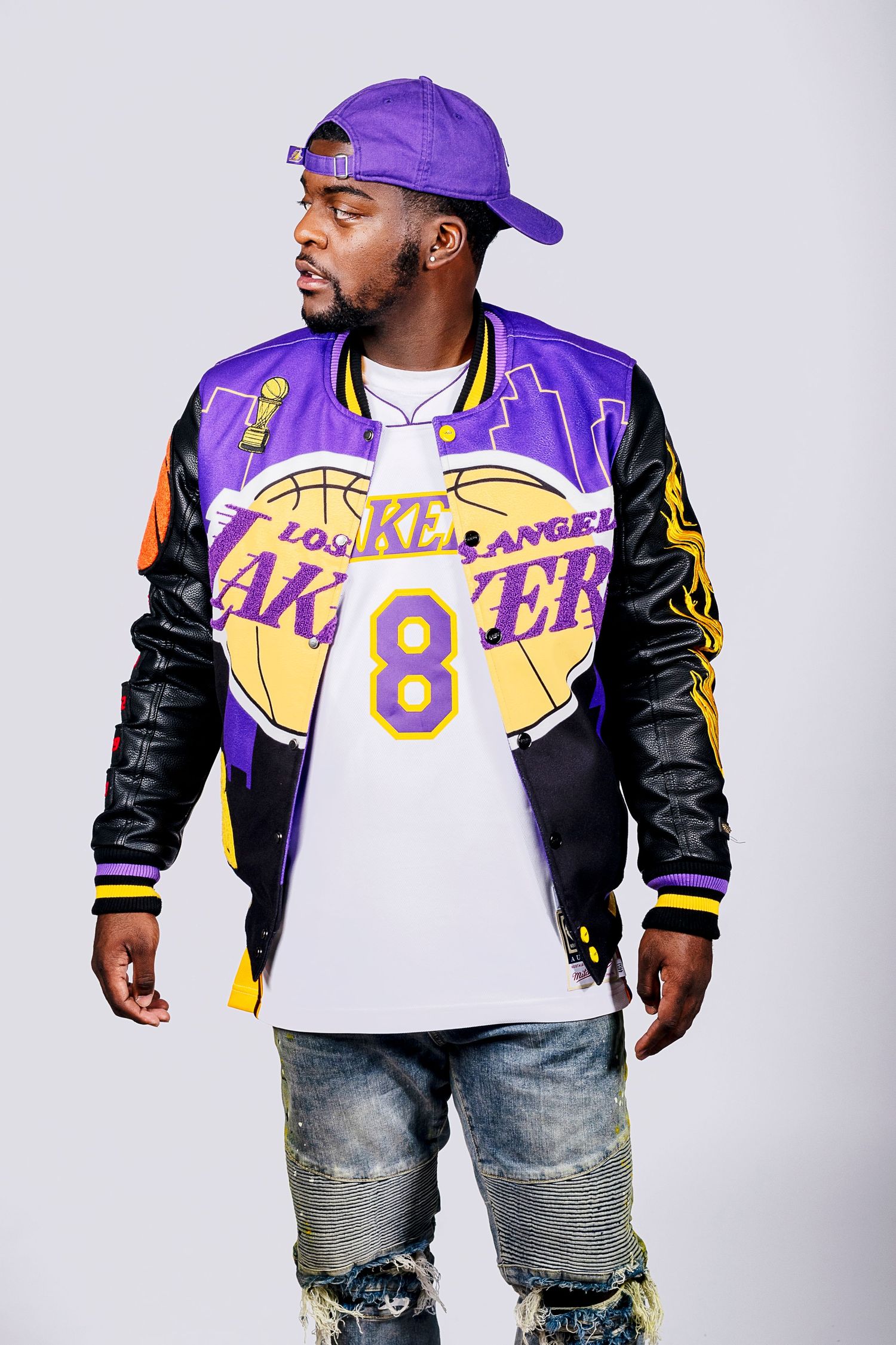 Man wearing a Los Angeles Lakers varsity jacket, purple cap, and ripped jeans posing confidently.