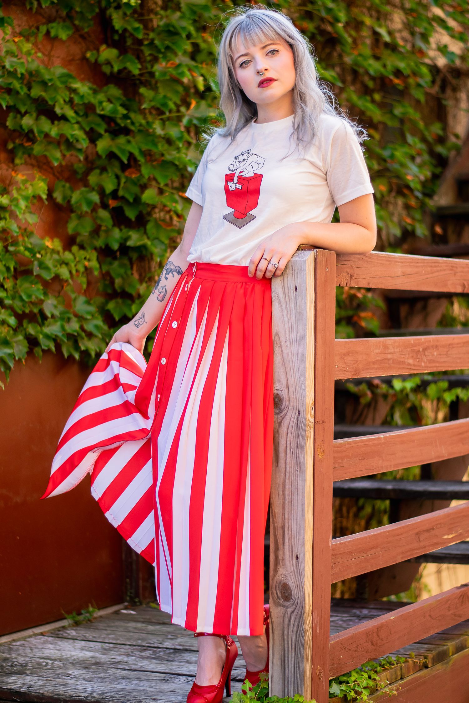 Young woman with silver hair wearing red striped skirt and white graphic tee leaning on wooden fence.