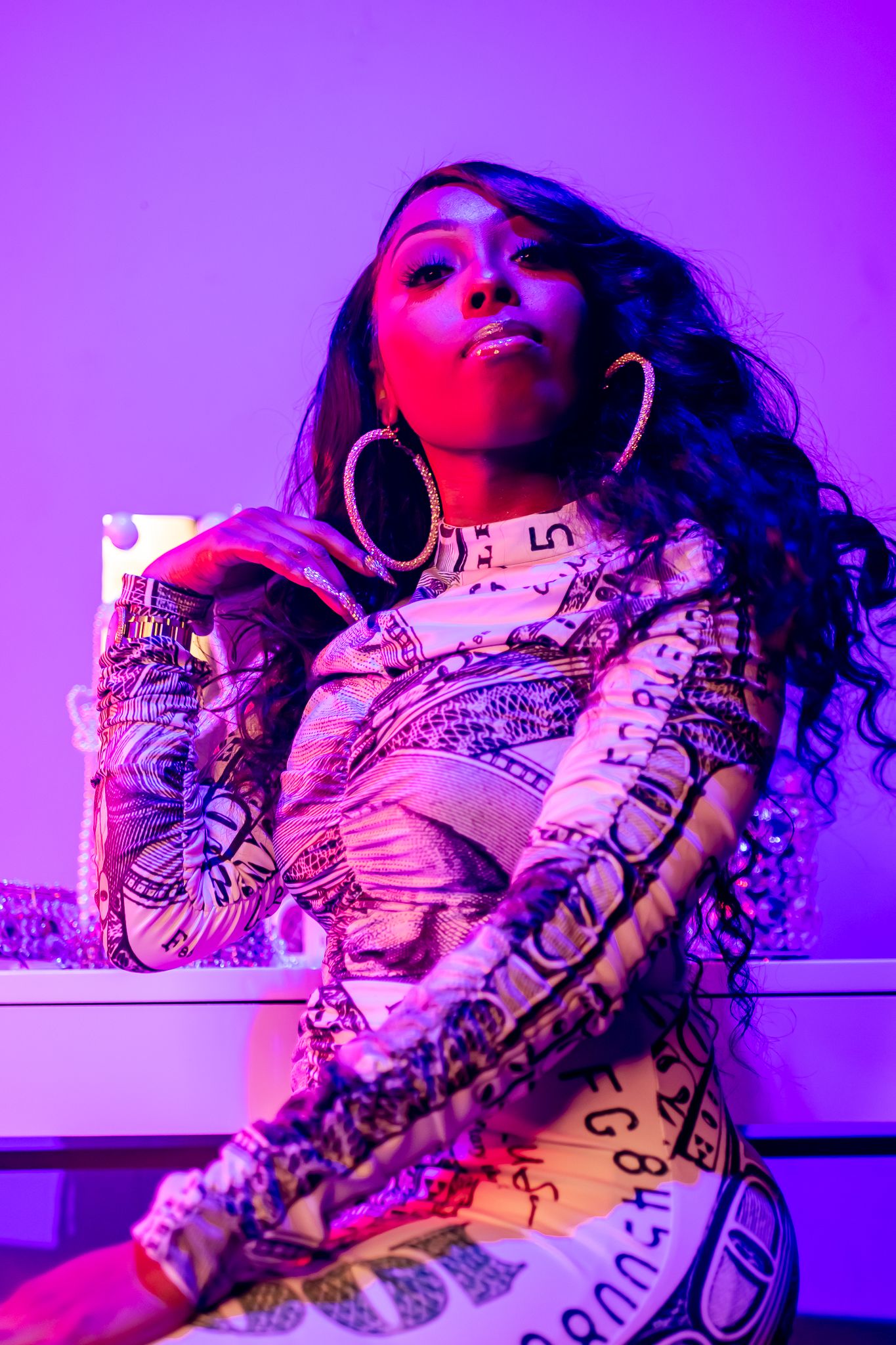 Woman in printed bodysuit posing under purple neon lighting with large hoop earrings.